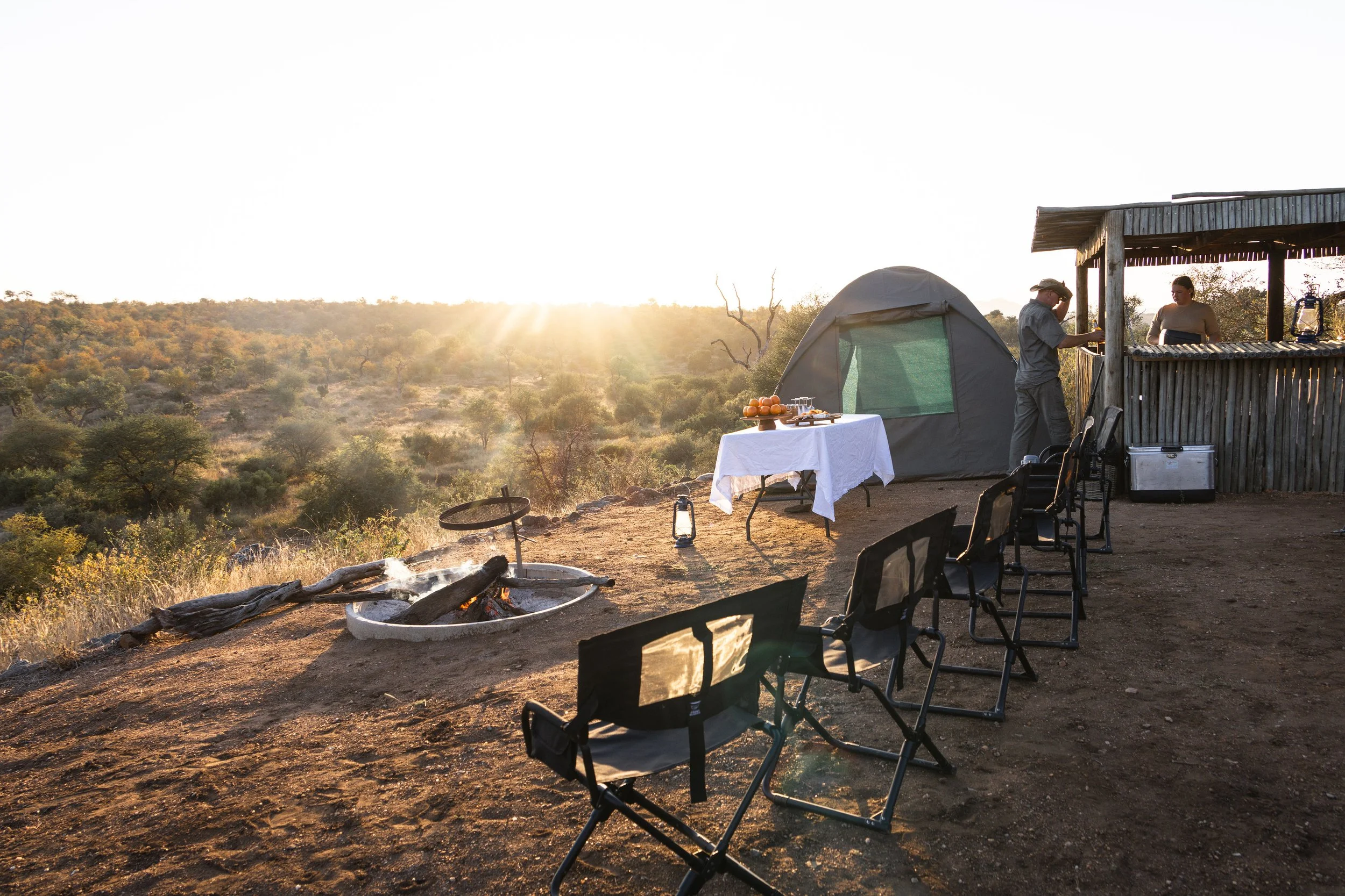Camping scene with a tent, a table with food, chairs, a fire pit, and two people at a rustic outdoor bar, overlooking a landscape at sunset.
