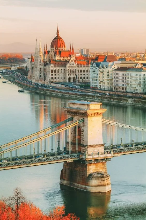 The Chain Bridge over the Danube River in Budapest, Hungary, with the Hungarian Parliament Building in the background during sunset.