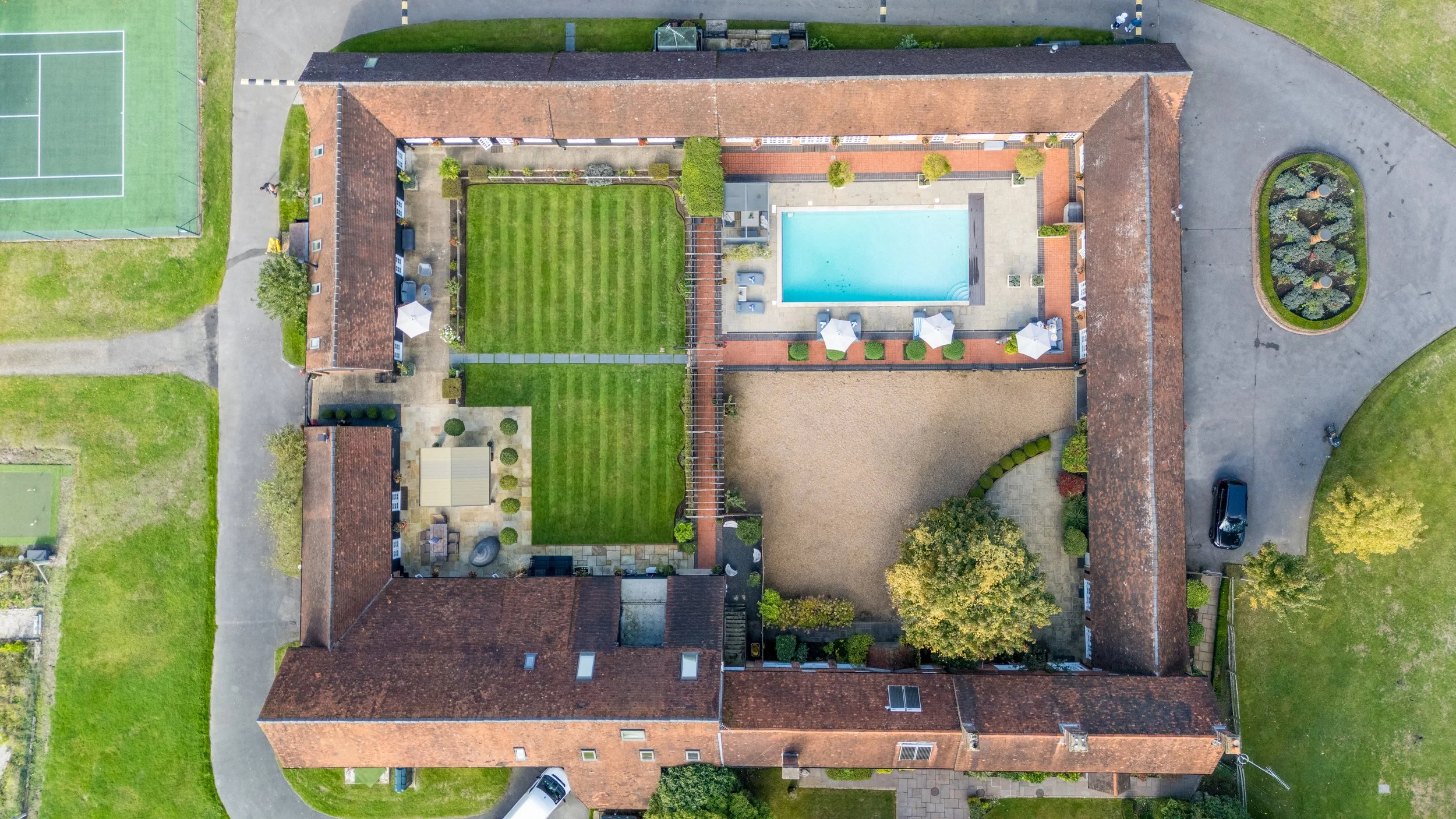 An aerial view of a large residential property showing a swimming pool, a tennis court, a landscaped garden, and surrounding buildings and pathways.