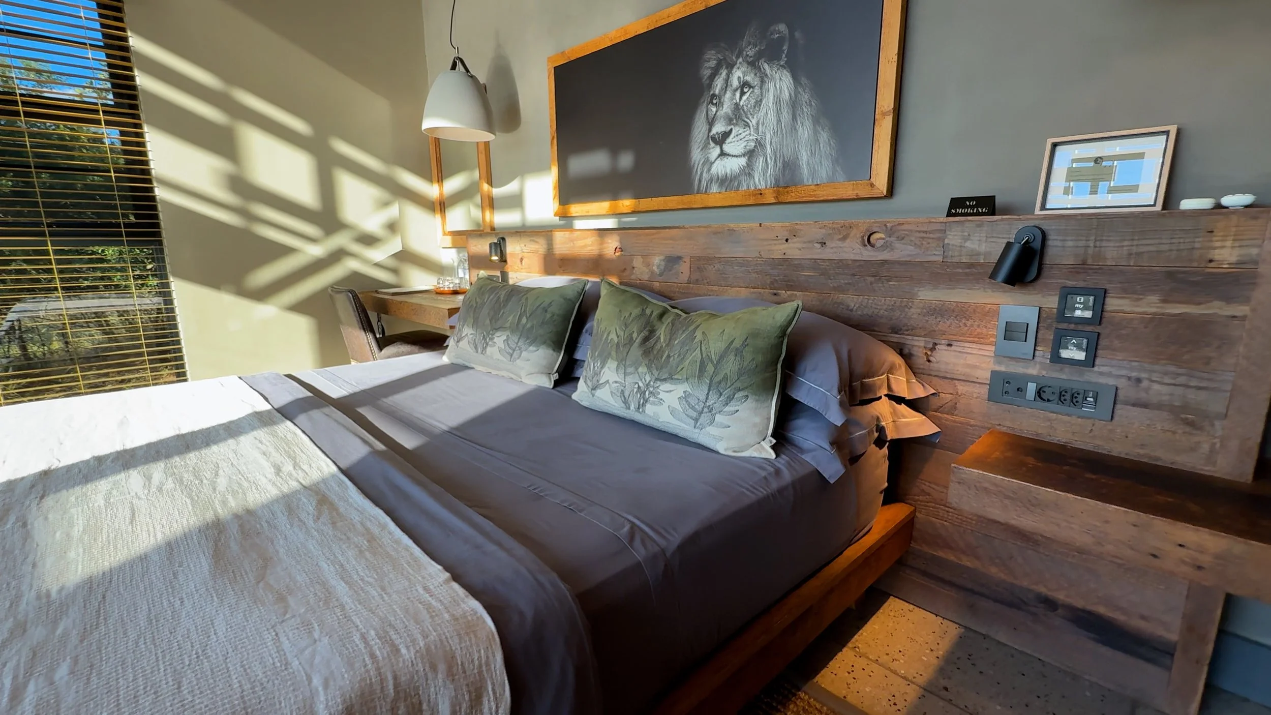Sunlight streaming through blinds onto a neatly made bed with green leaf-patterned pillows in a modern rustic bedroom with wooden accents and wall art.