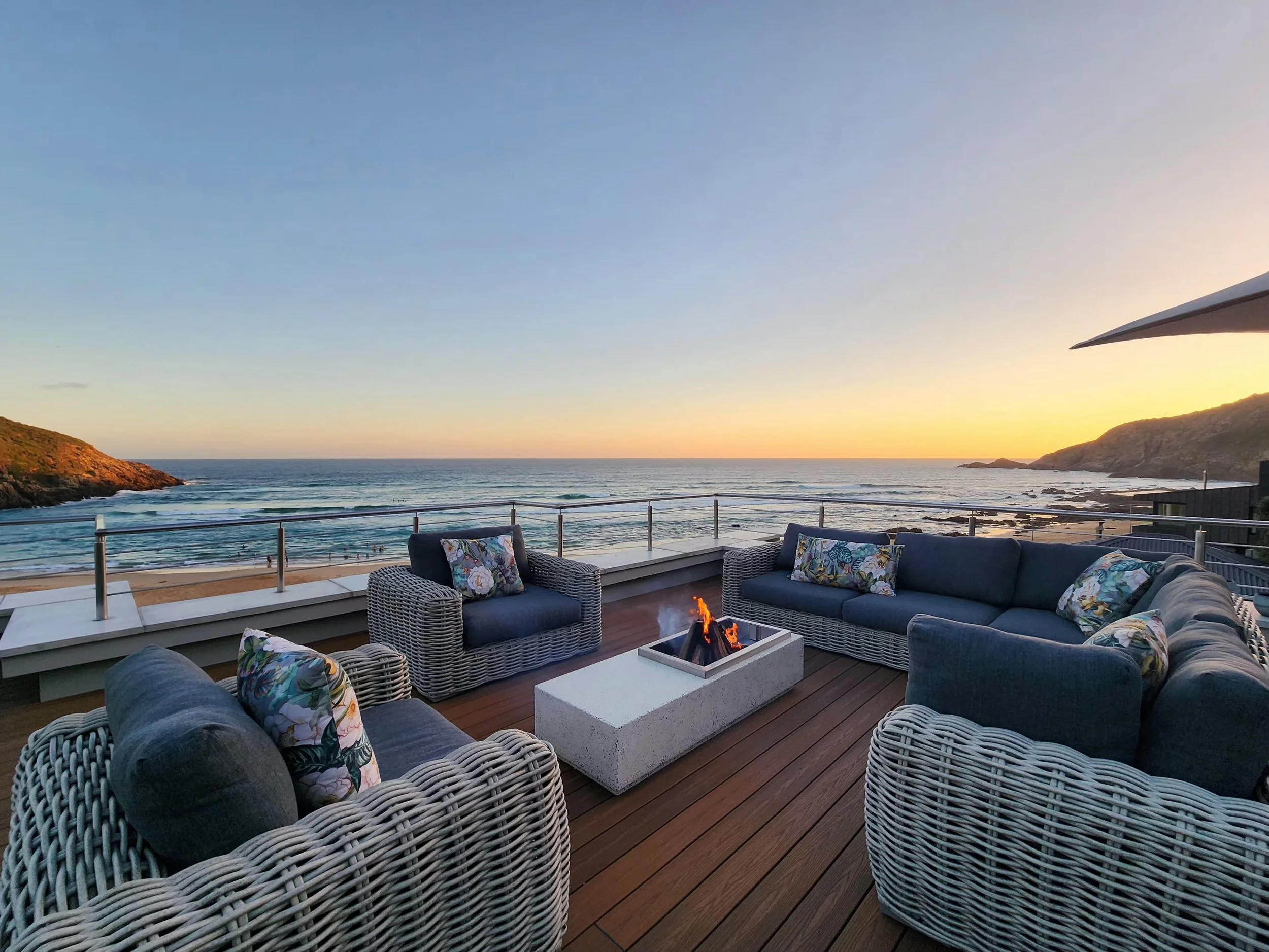 Outdoor patio with wicker and cushioned furniture arranged around a small fire pit, overlooking the ocean at sunset with hills in the background.