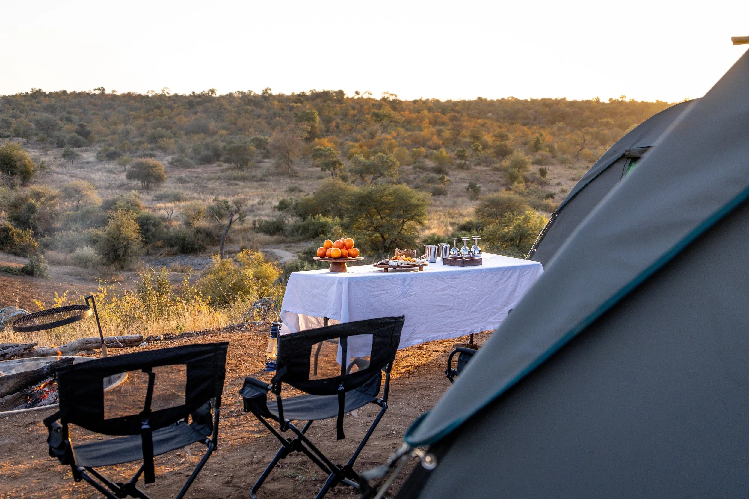 Camping scene with a table set with oranges, snacks, and glasses behind tents overlooking a hilly landscape with trees during sunset.
