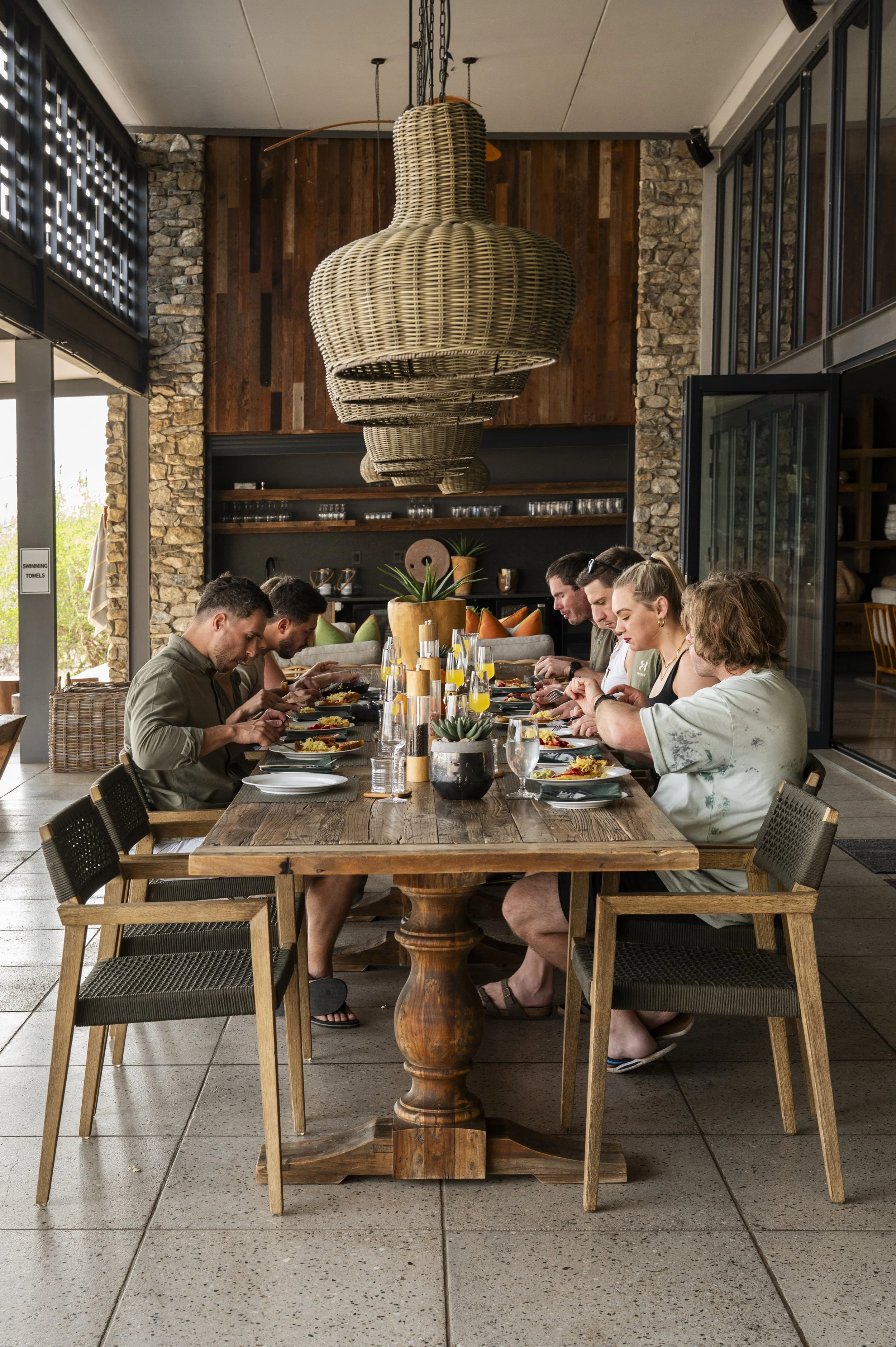 Group of six people sitting at a long wooden table, eating a meal together in a restaurant with modern decor and large baskets hanging from the ceiling.