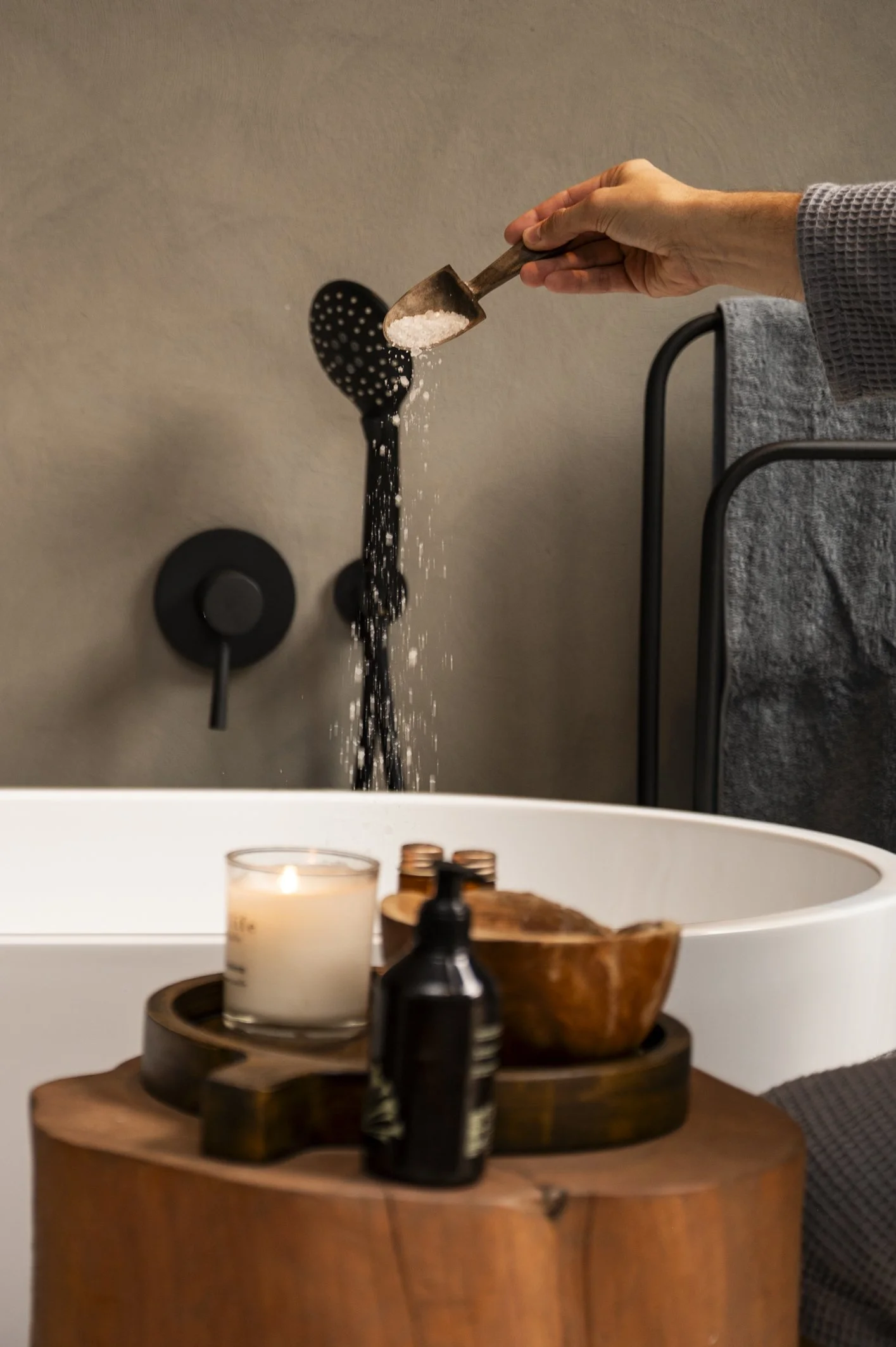 A person pours pink bath salts from a small wooden scoop into a black slotted spoon placed in a bath. The scene includes a candle, a black lotion pump, and a wooden bowl on a wooden tray set on a side table next to a white bathtub.
