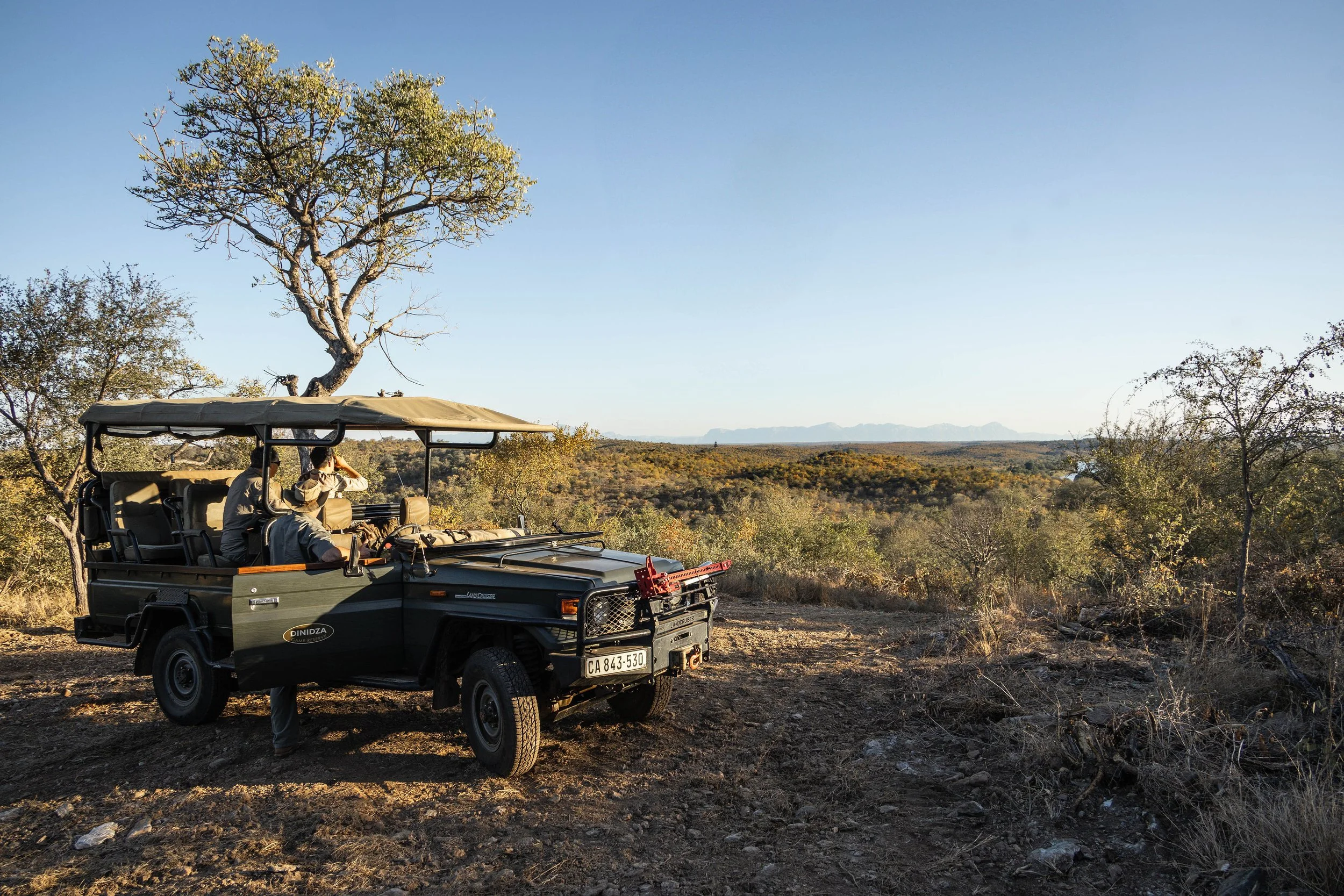A safari vehicle with three people inside, parked in a dry, open landscape surrounded by sparse trees and distant mountains in the background, during daylight.