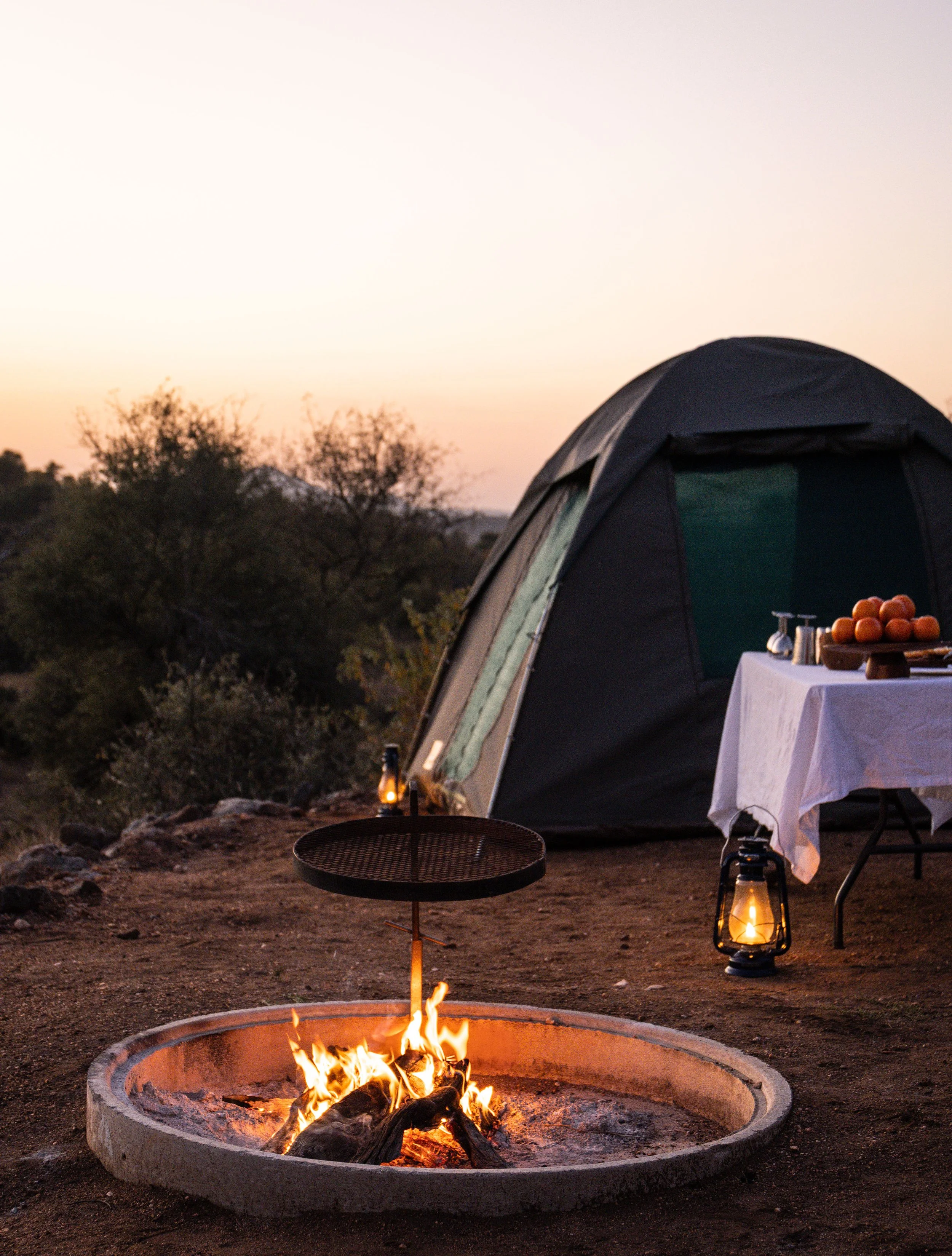 Camping scene with a tent, a campfire, a table with food, and lanterns at sunset in a natural outdoor setting.