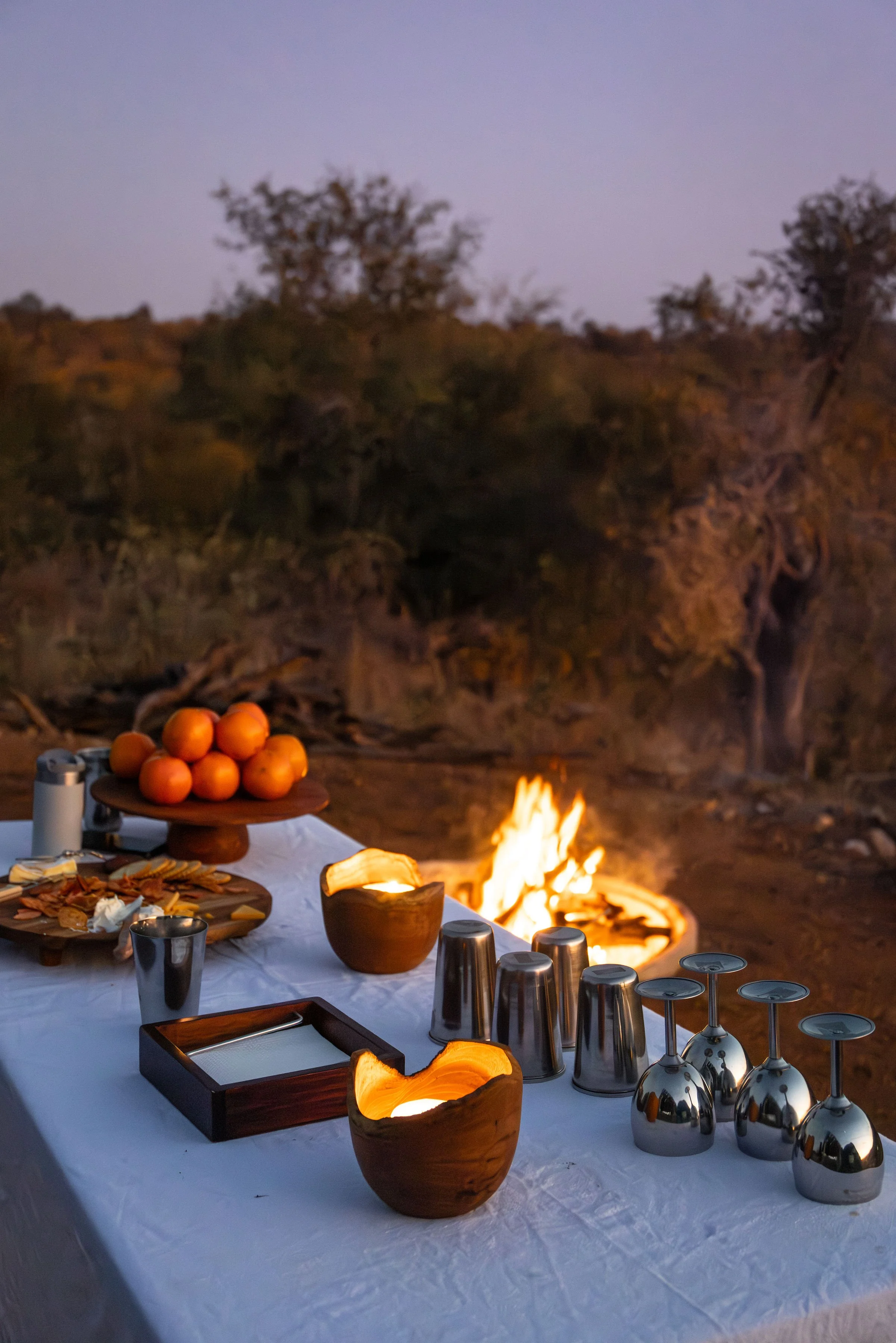 Outdoor dining setup with a white tablecloth, candles, metal cups, glassware, a wooden bowl, a tray, and a large bowl of oranges with a campfire burning in the background.