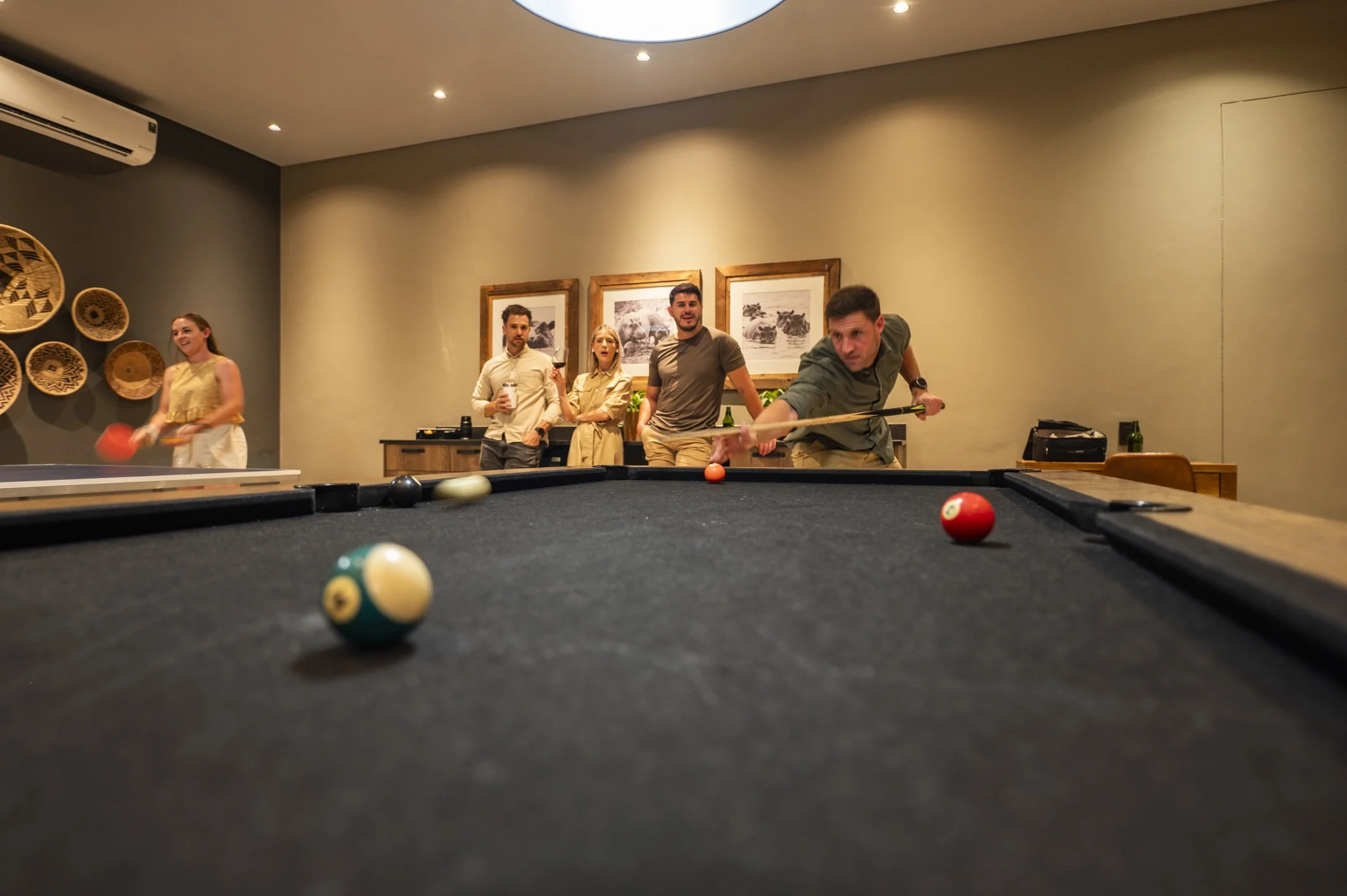 Group of five people playing pool in a game room, with one person taking a shot and others watching, in a room decorated with framed photographs and wall baskets.