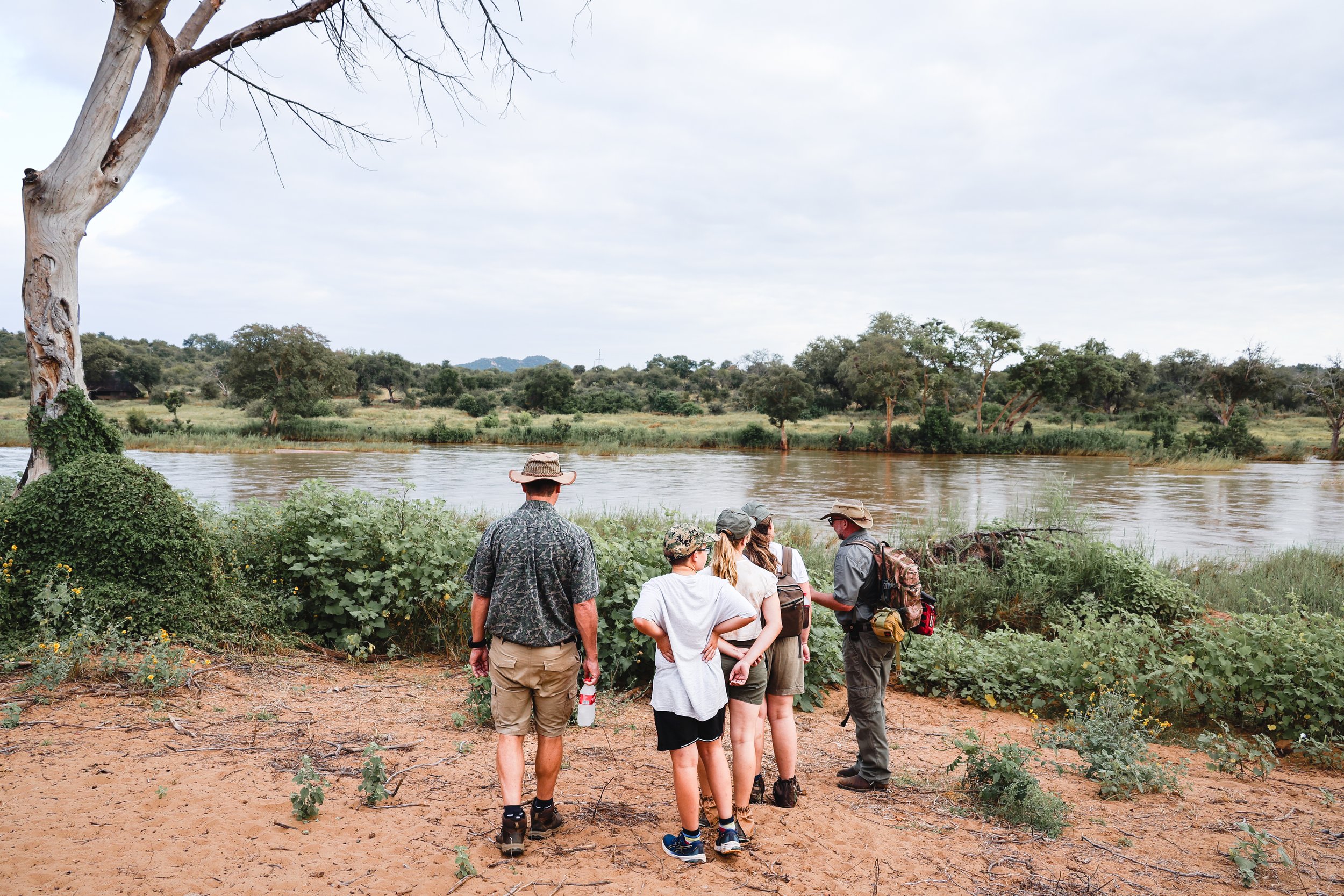 Group of people on a nature walk by a river, surrounded by greenery and trees, with cloudy sky overhead.