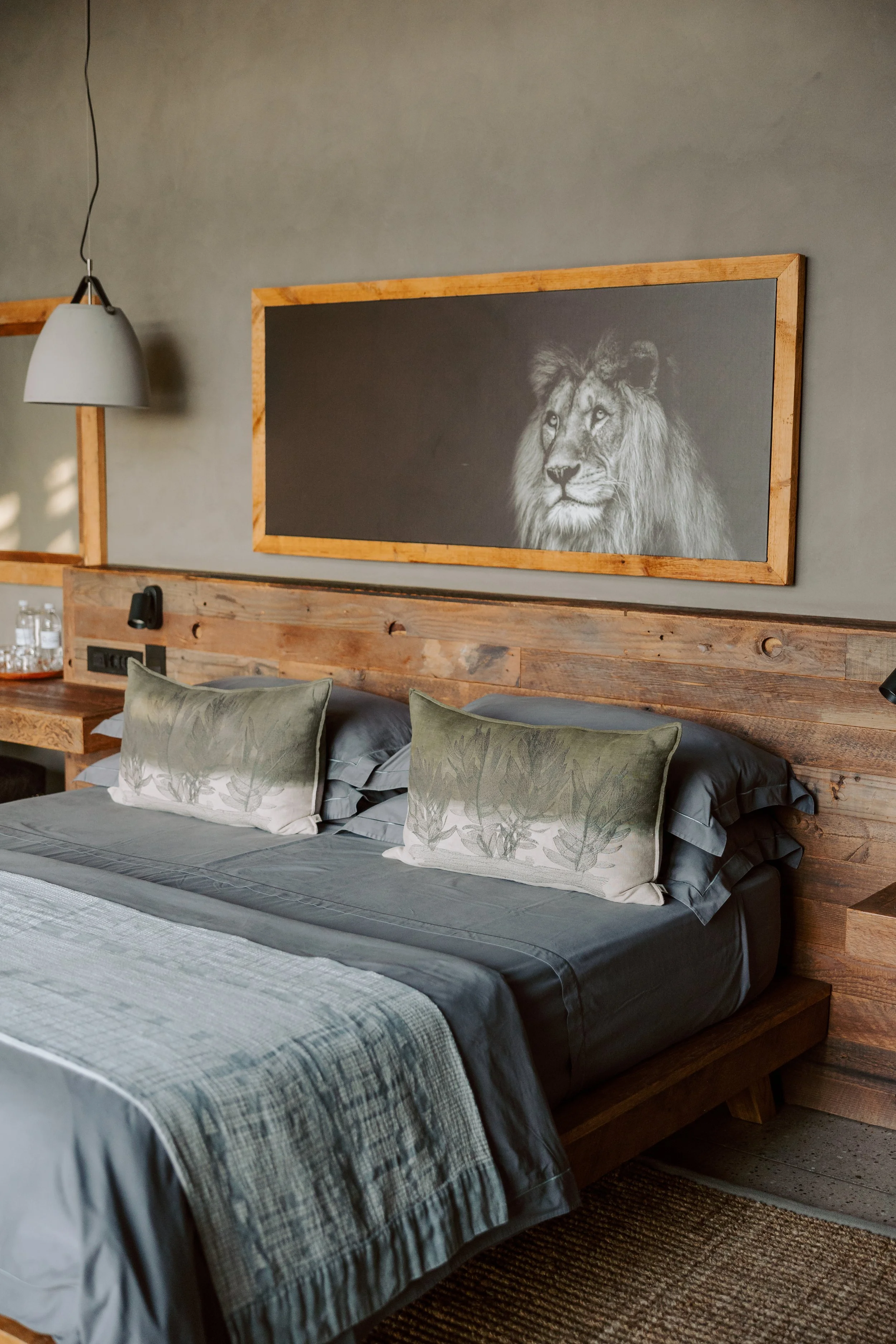 A bedroom with a wooden headboard, two green and white pillows, gray bedding, a black and white lion artwork above the bed, a wall-mounted lamp, and a small wooden nightstand with water bottles.