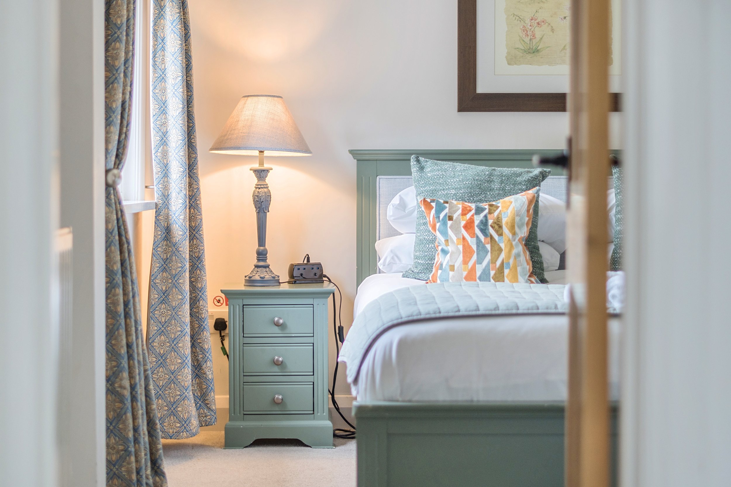 A cozy bedroom with a green headboard, patterned pillows, and a white bedspread. A nightstand with a lamp and a clock, curtains with a blue and gold pattern, and framed art on the wall.