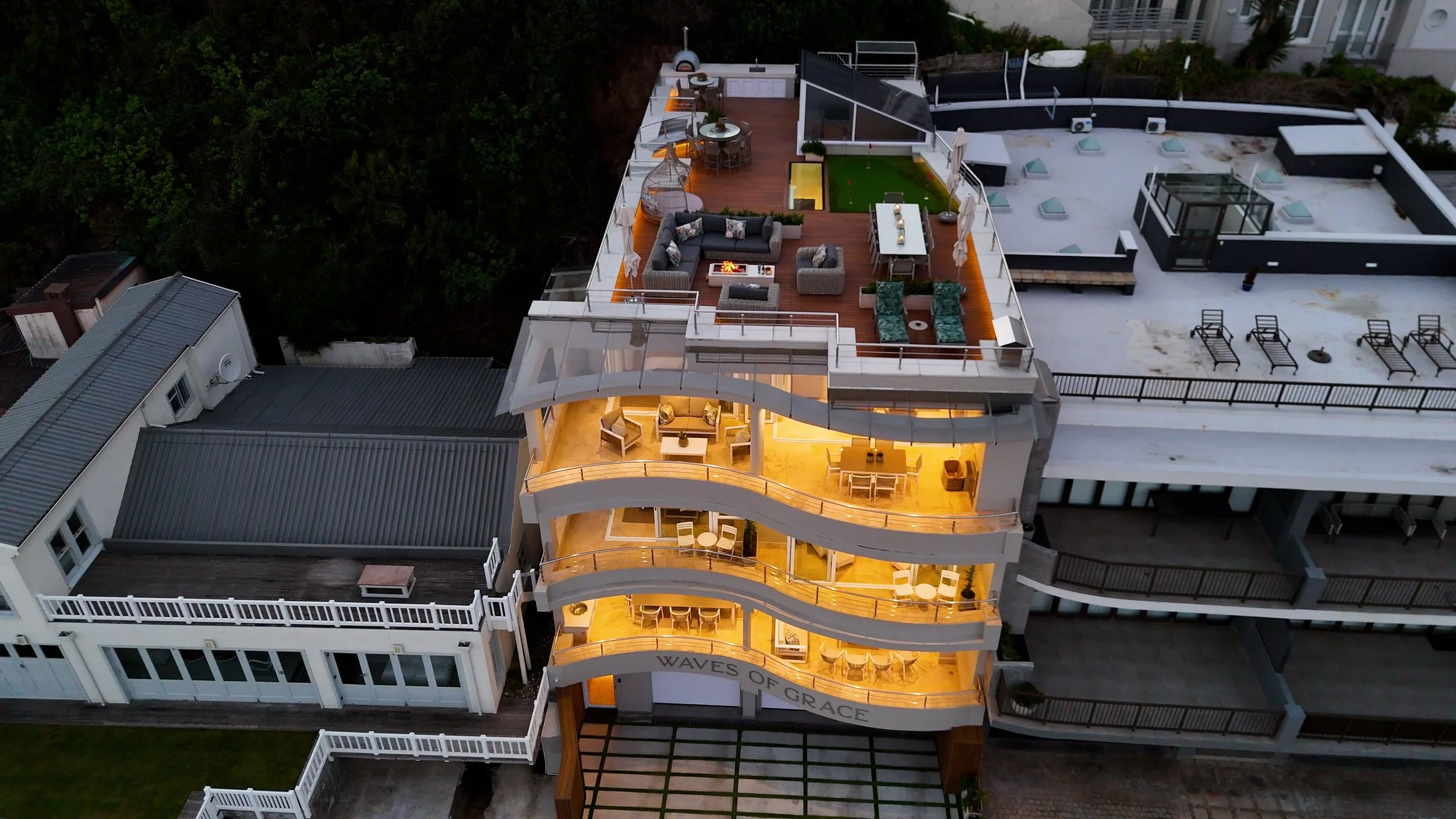 Aerial view of a rooftop apartment building with illuminated balconies and a furnished outdoor terrace with seating, a fire pit, and a small enclosed green area.