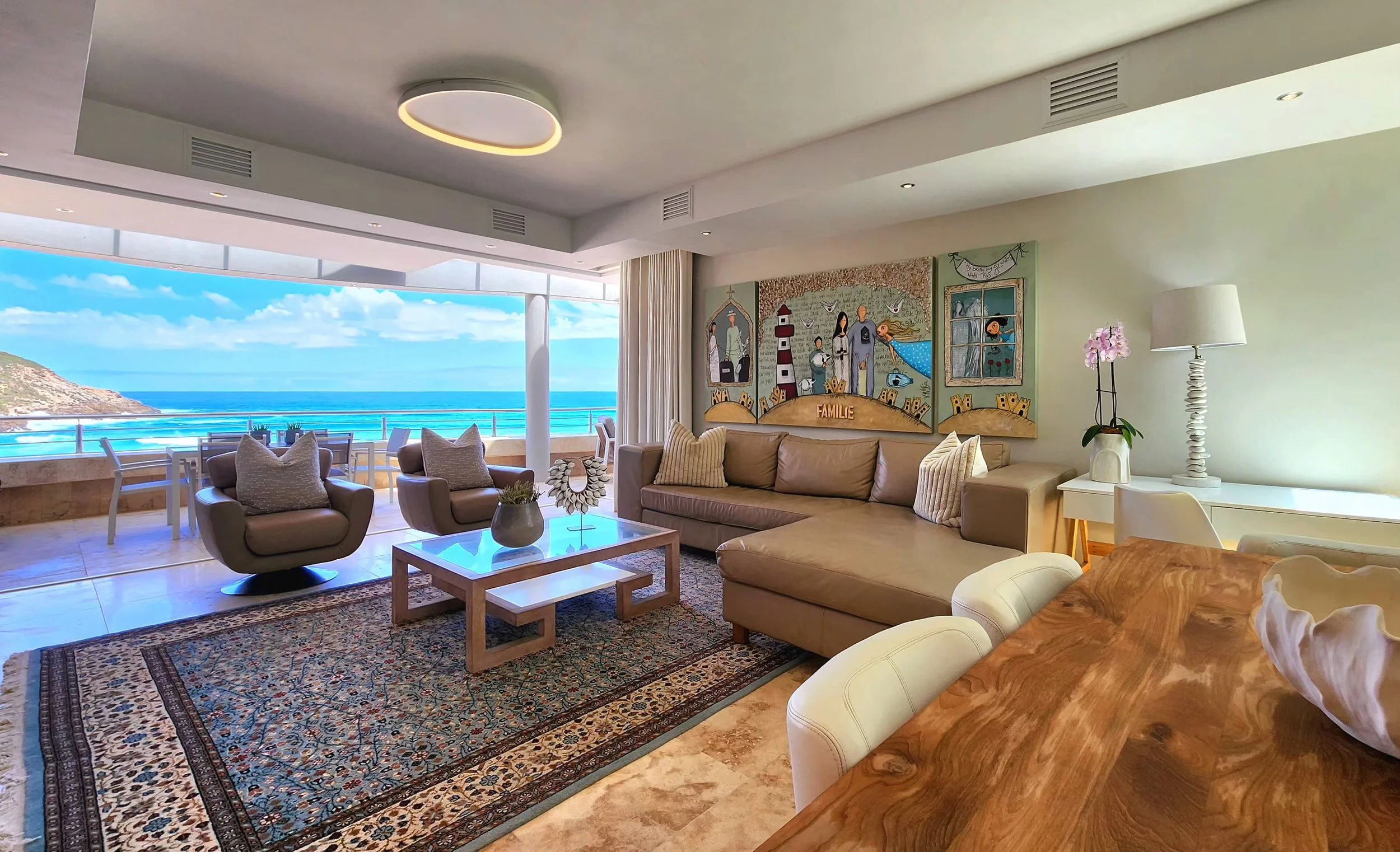 Living room with ocean view, beige sofa, armchairs, coffee table, patterned rug, wall art, and balcony with outdoor seating.