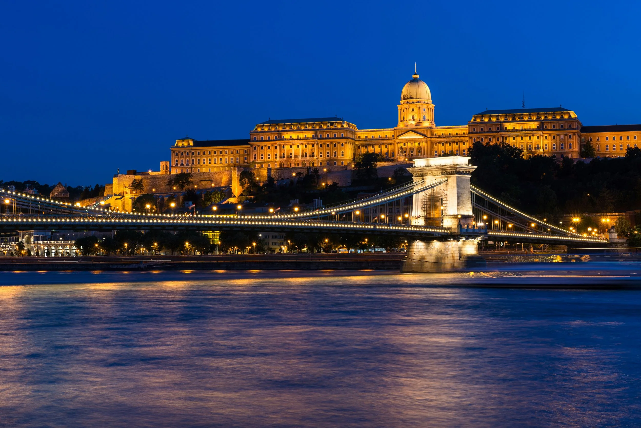 Night view of a large, illuminated historic building with a dome, situated on a hill, with a suspension bridge lit up in the foreground over a river.