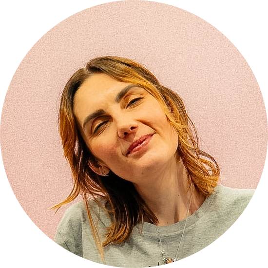 Amy with shoulder-length light brown hair and a grey t-shirt, smiling, against a pink background.