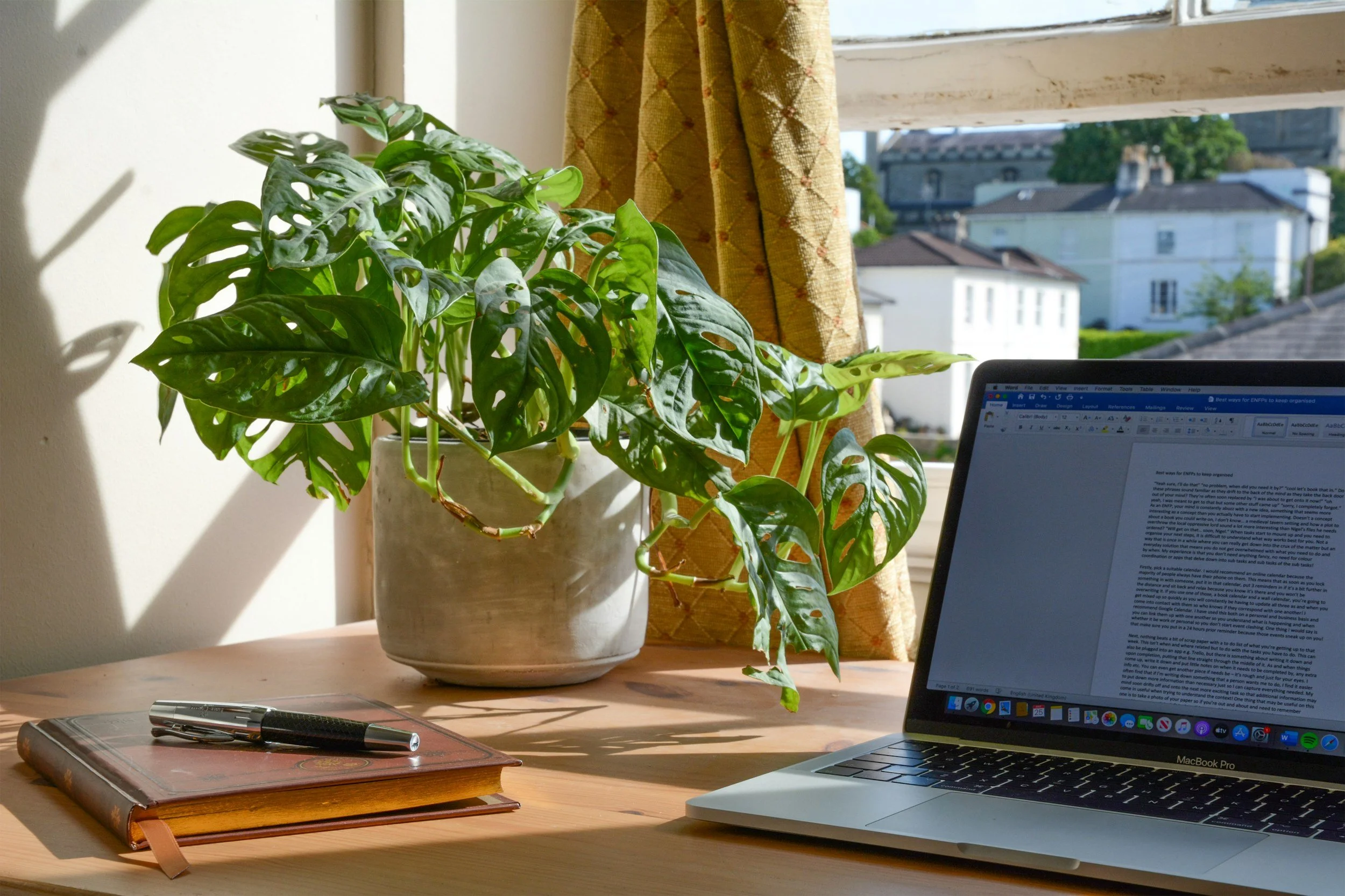 A laptop open on a wooden desk next to a brown notebook with a black pen on top. A large potted Monstera plant is beside the laptop. Sunlight streams through a window, casting shadows on the desk, with a view of buildings outside.