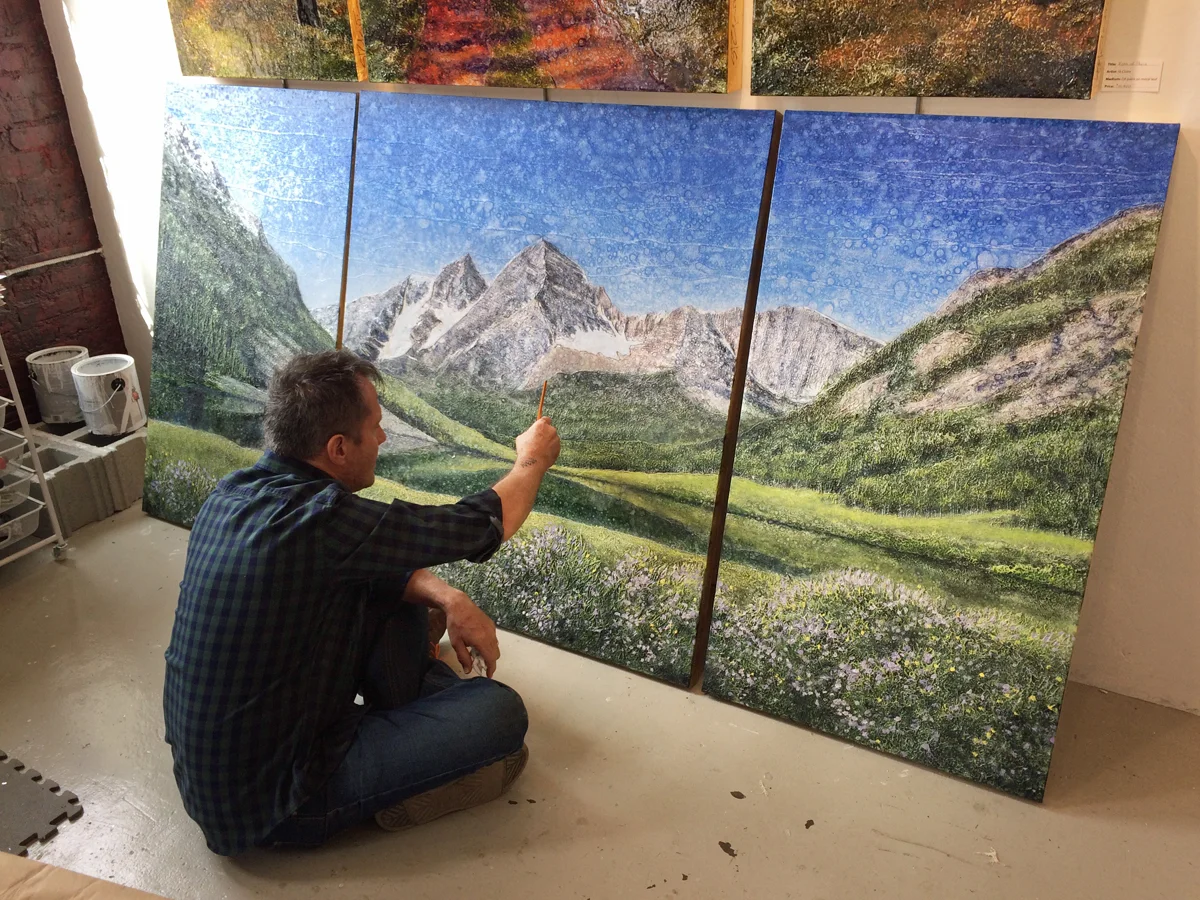 Oil painter Stephen St. Claire sitting on the floor with a paintbrush, working on a three-panel landscape painting of mountains, green fields, and a blue sky with clouds.