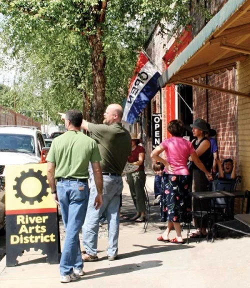 People standing outside a store with a red, white, and blue flag, a sign for River Arts District, and an open sign on the door, with trees and buildings in the background.