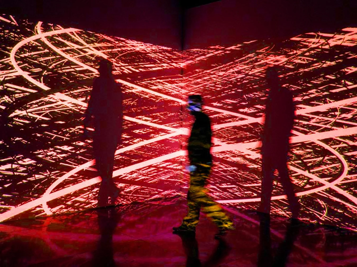 Three silhouetted figures inside a dark immersive art installation, the walls and floor lit with swirling red and pink projected lines. One figure is mid-stride, captured in motion blur.