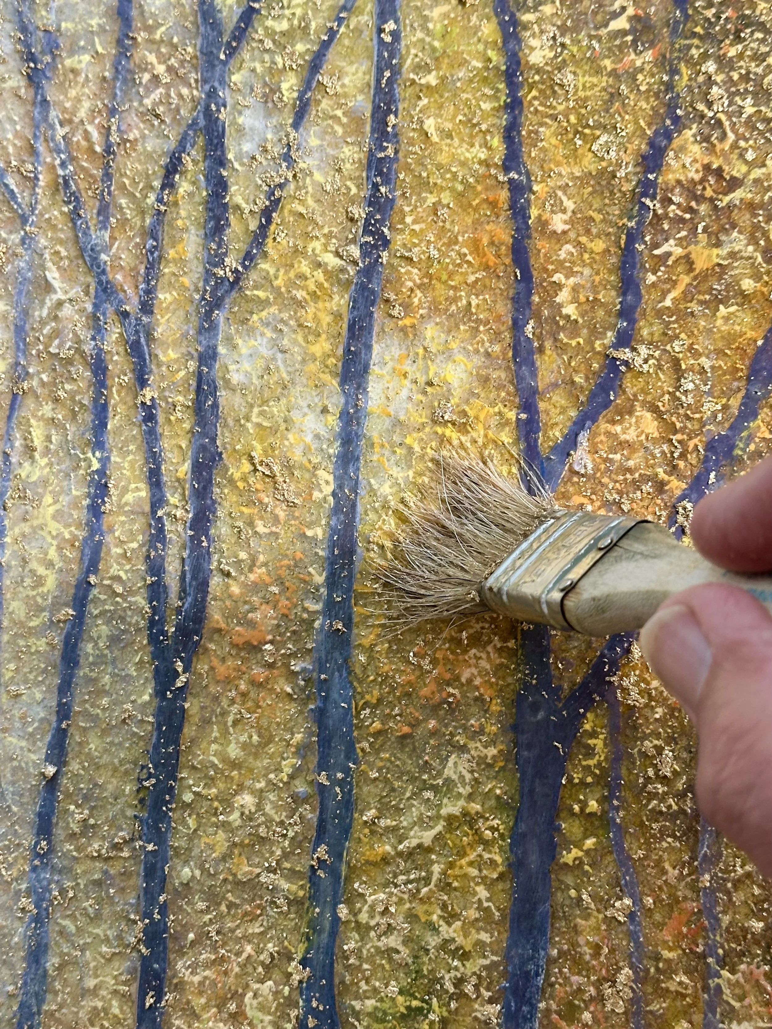 A hand holding a paintbrush painting tree branches on a textured, abstract background with streaks of blue and yellow, resembling a natural landscape.