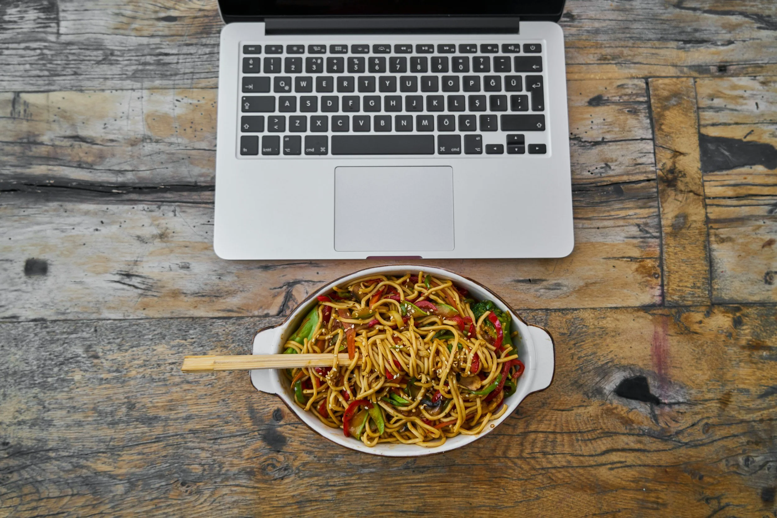 A laptop and a bowl of stir-fried noodles with vegetables on a rustic wooden table.