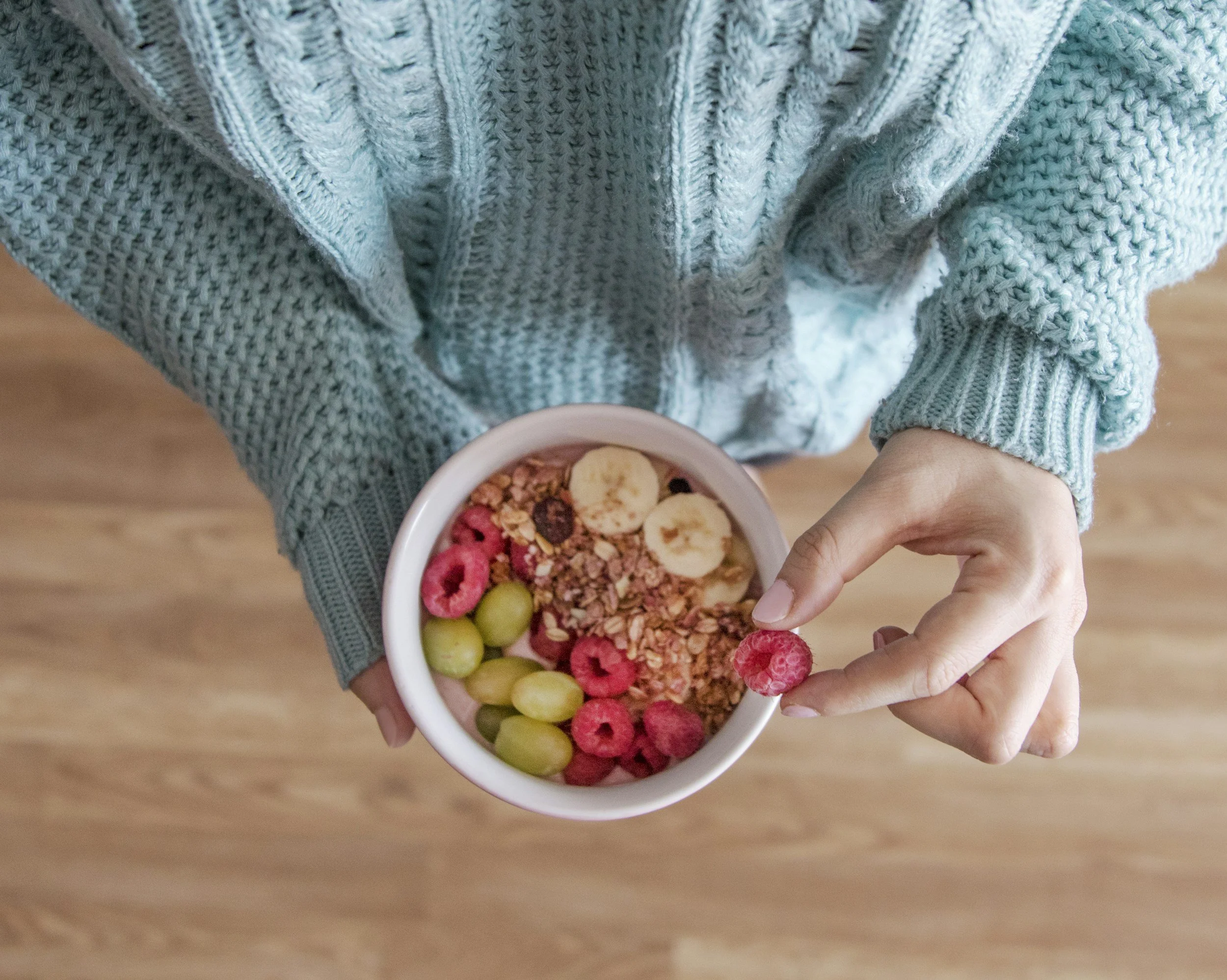 A person wearing a blue cable-knit sweater holding a bowl of cereal with bananas, raspberries, grapes, and granola. The person is picking up a raspberry with their hand.
