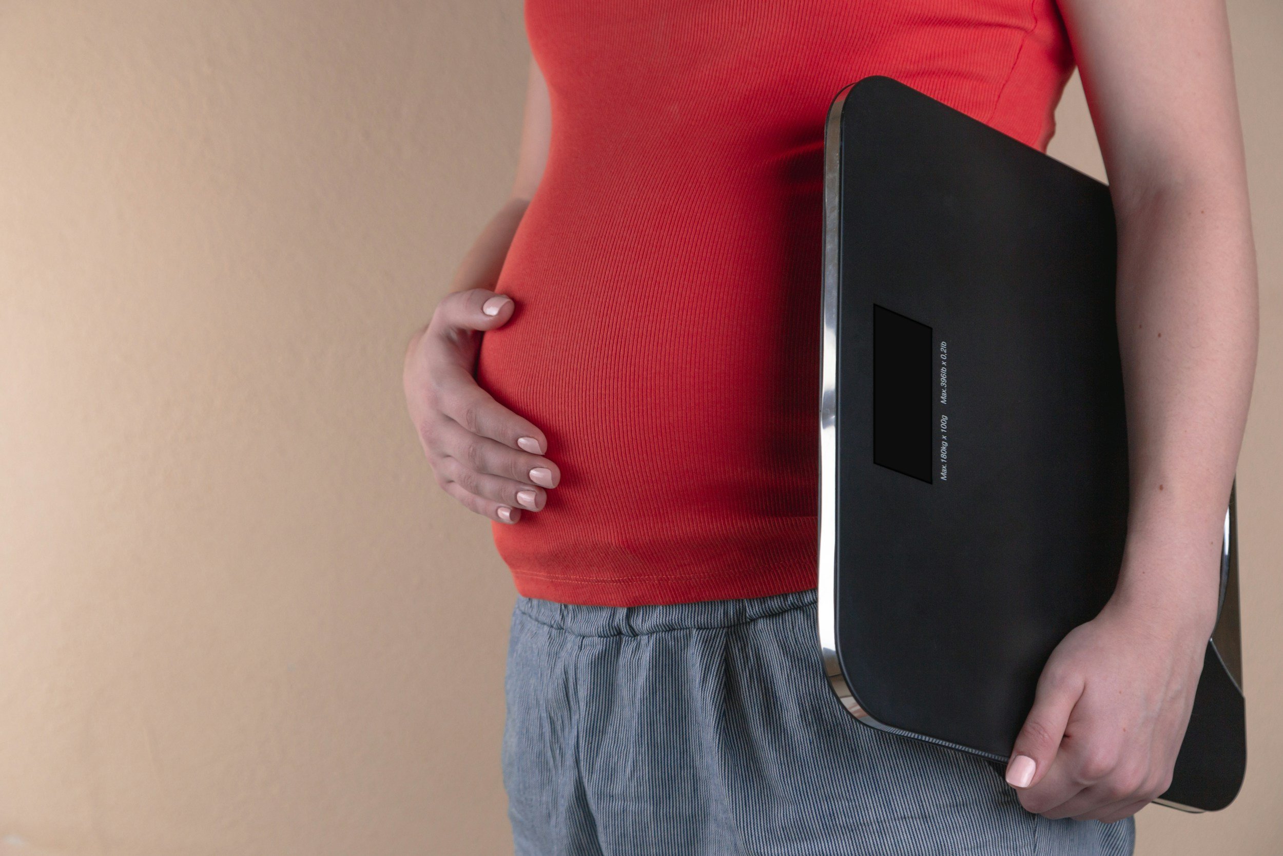 A pregnant woman in a red shirt holding her belly with one hand and carrying a black folder or laptop under her other arm, standing against a plain beige background.