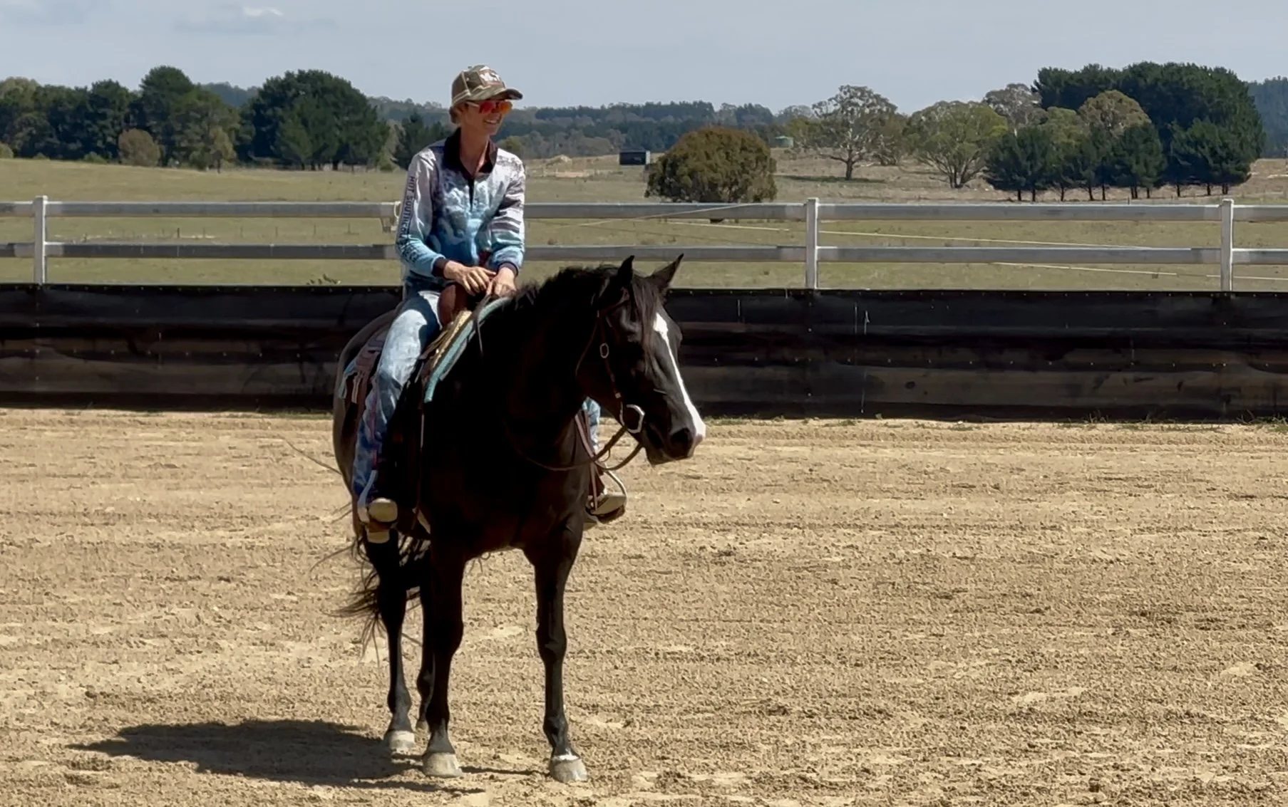 A woman wearing a cowboy hat and sunglasses riding a black and white horse on a dirt arena outdoors, with trees and blue sky in the background.