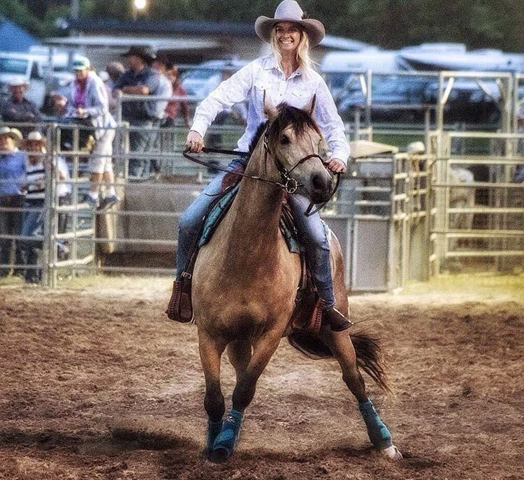 Woman in cowboy hat riding a brown horse inside a rodeo arena with onlookers and vehicles in the background.