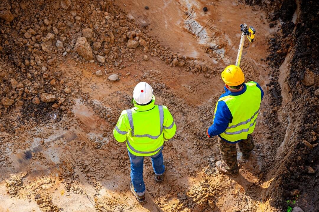 Two geotechnical engineers in high-visibility jackets and helmets assessing exposed soil layers during a geological assessment on a construction site.