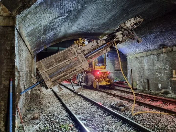 Rail-mounted drilling rig installing ground anchors into a brick retaining wall during a Phase 2 geotechnical investigation.