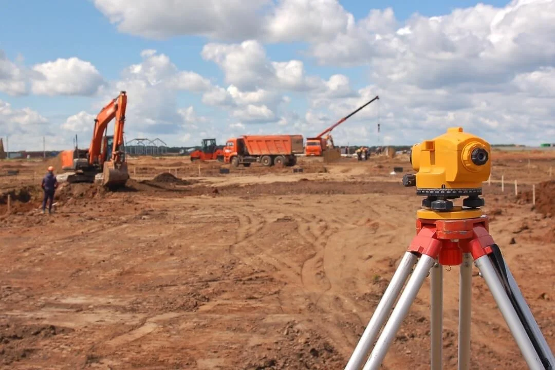 Engineering level on tripod overlooking excavators, dump truck and crane during a site walkover survey to plan ground investigation.
