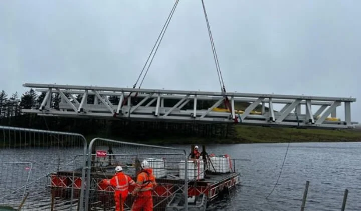 Engineers installing a steel bridge structure over Penwhapple Reservoir using a crane and floating platform.