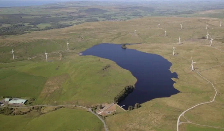 Aerial view of Penwhapple Reservoir surrounded by green hills and wind turbines in the landscape.