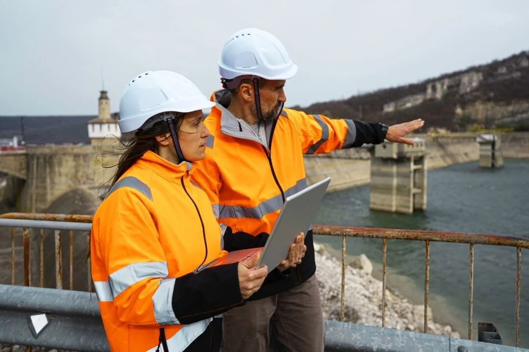 Geotechnical engineers performing a site walkover survey at a dam, recording ground conditions with a laptop.
