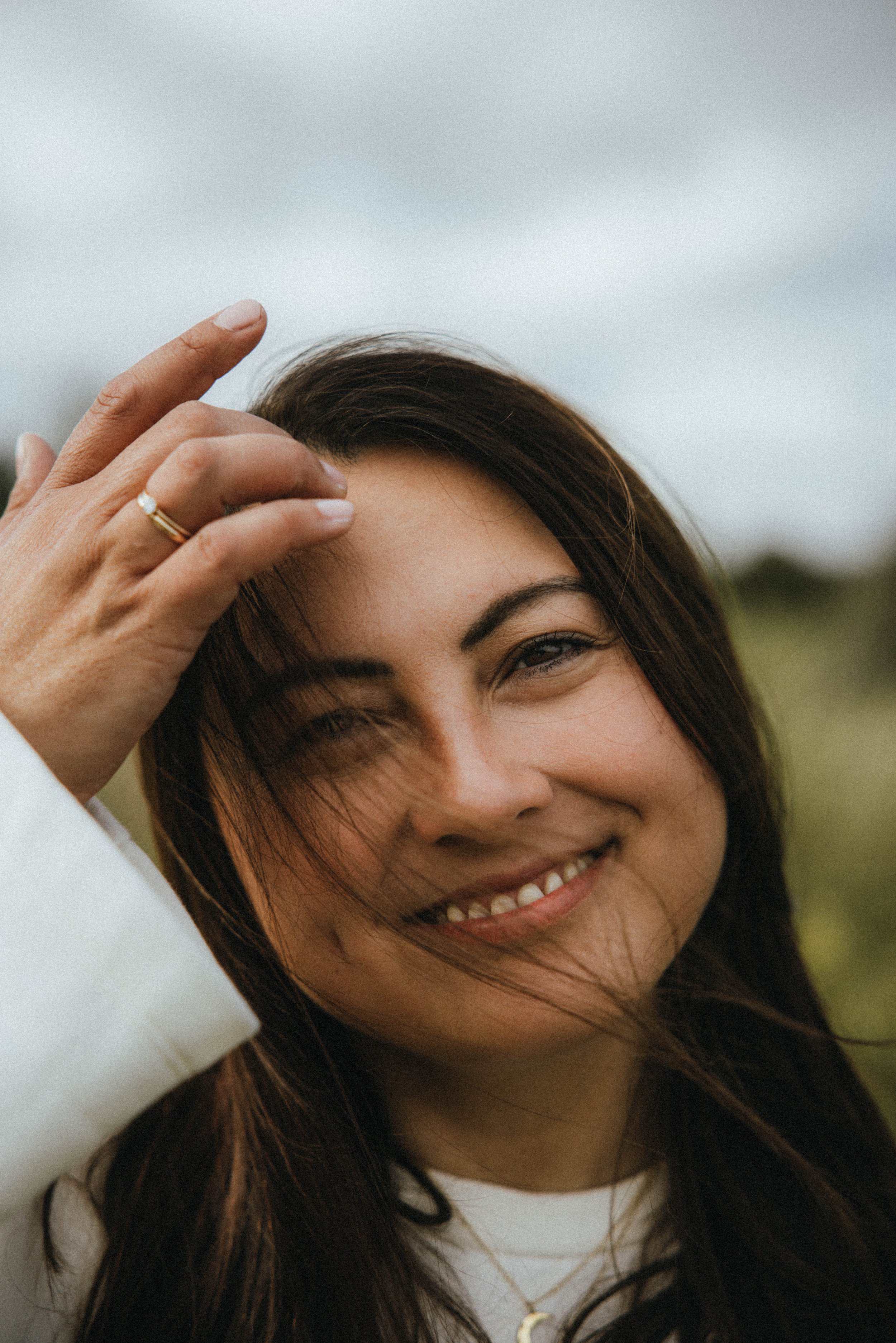 Lächelnde Frau mit dunklen Haaren, die im Freien steht, die Hand auf der Stirn, gegen den Wind, bei bewölktem Himmel.