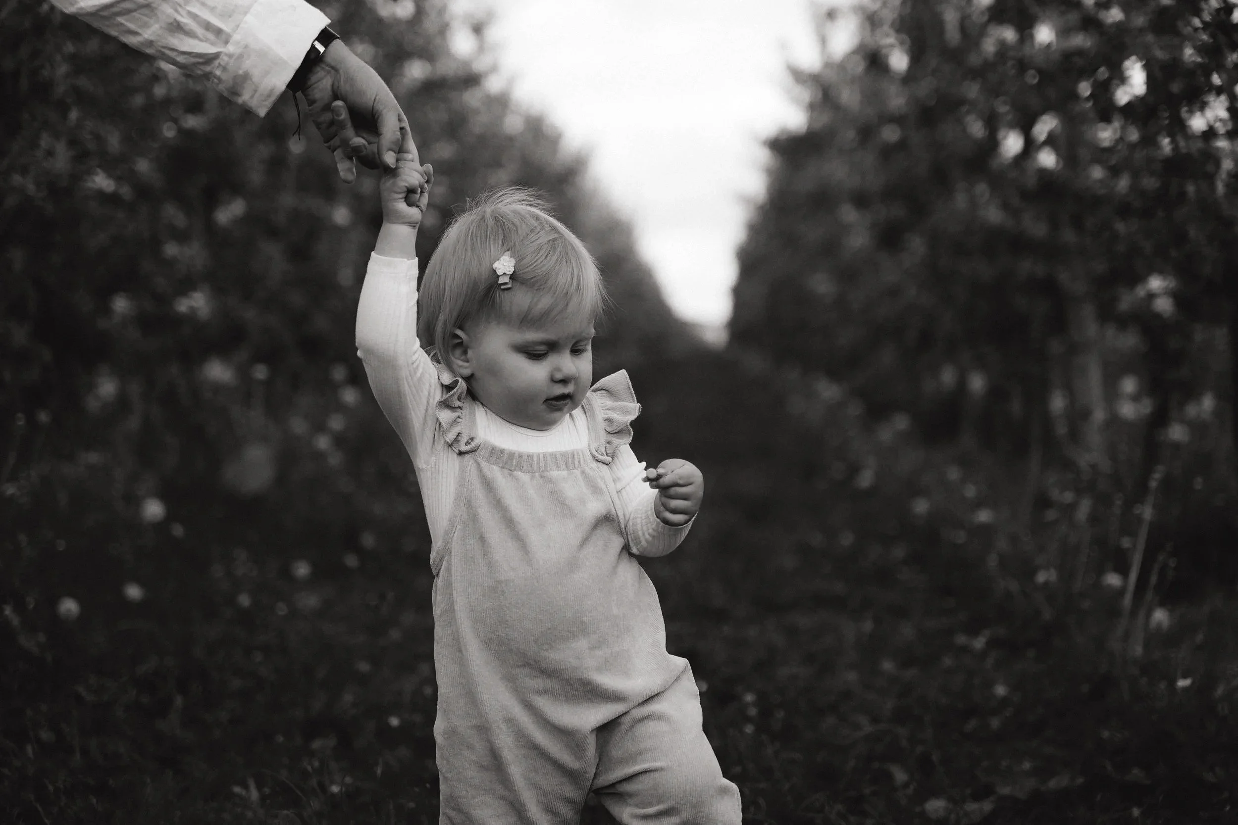 Ein kleines Mädchen in einem Outdoor-Umfeld, das von einer erwachsenen Hand gehalten wird, während es auf dem Boden steht.