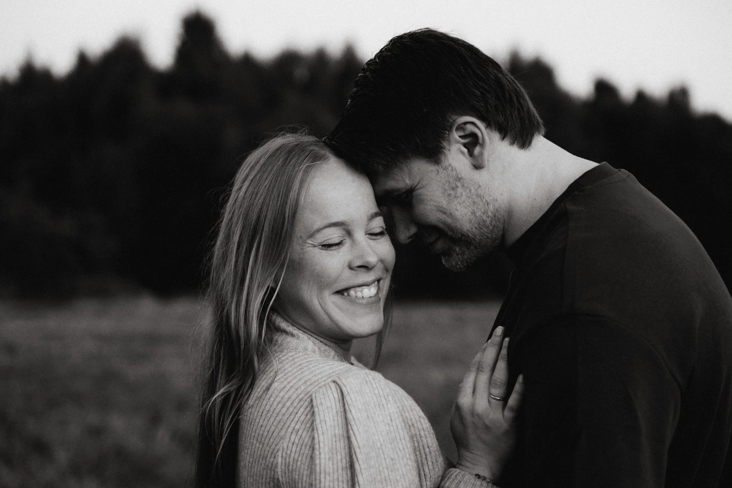 Ein lachendes Paar steht eng beieinander im Freien, Kopfhäuser berühren sich, im Hintergrund Bäume, schwarz-weiß Foto.