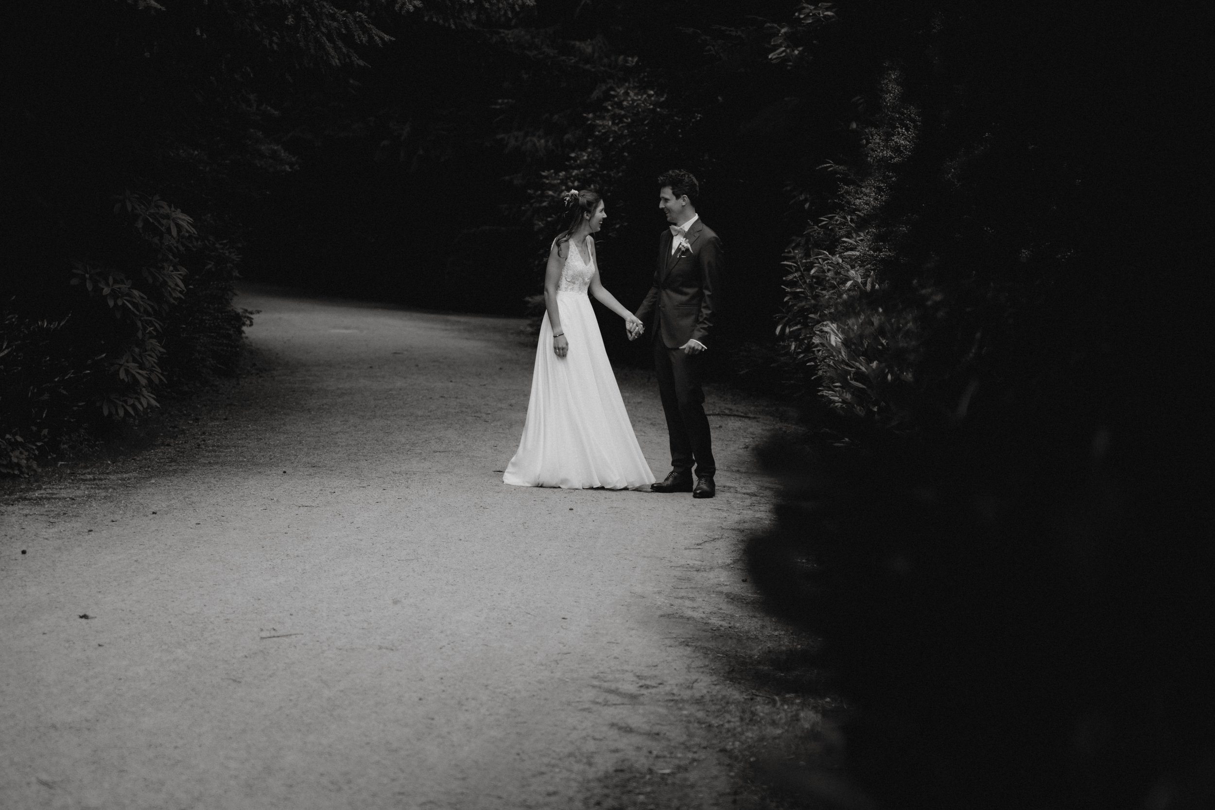 Ein Brautpaar hält sich an einem dunklen Waldweg bei Nacht an den Händen. Sie tragen Hochzeitskleider und sehen sich verliebt an.
