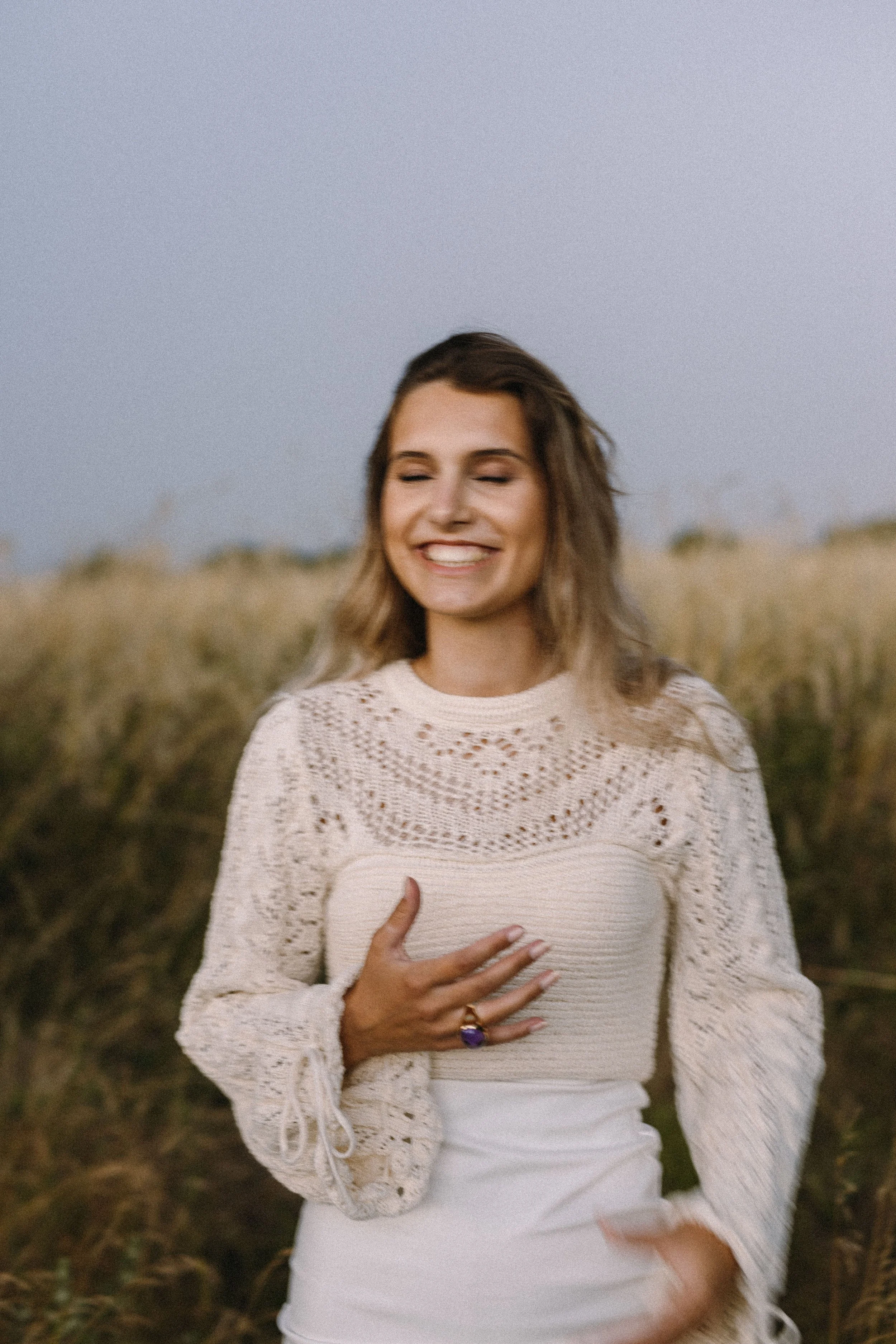 Eine lachende Frau im Freien, trage ein weißes, handgestricktes Oberteil mit offenen Mustern, in einem Feld mit hohen Gräsern, bei Sonnenuntergang.