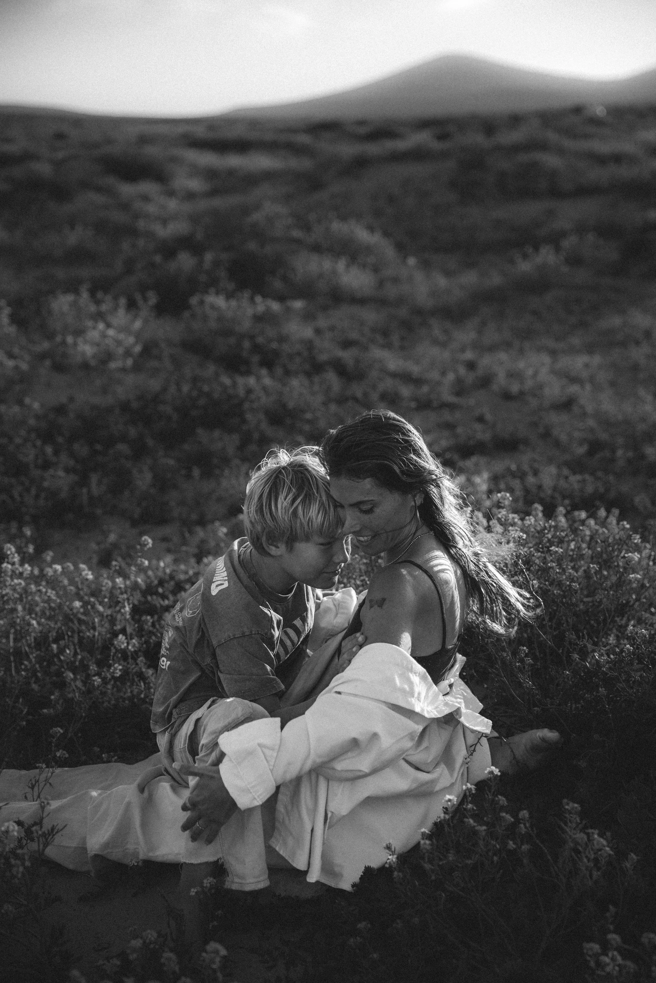 Eine Frau und ein Kind sitzen in einem Feld mit Blumen, eng umschlungen und lächelnd, bei Sonnenuntergang, schwarz-weiß Fotografie.