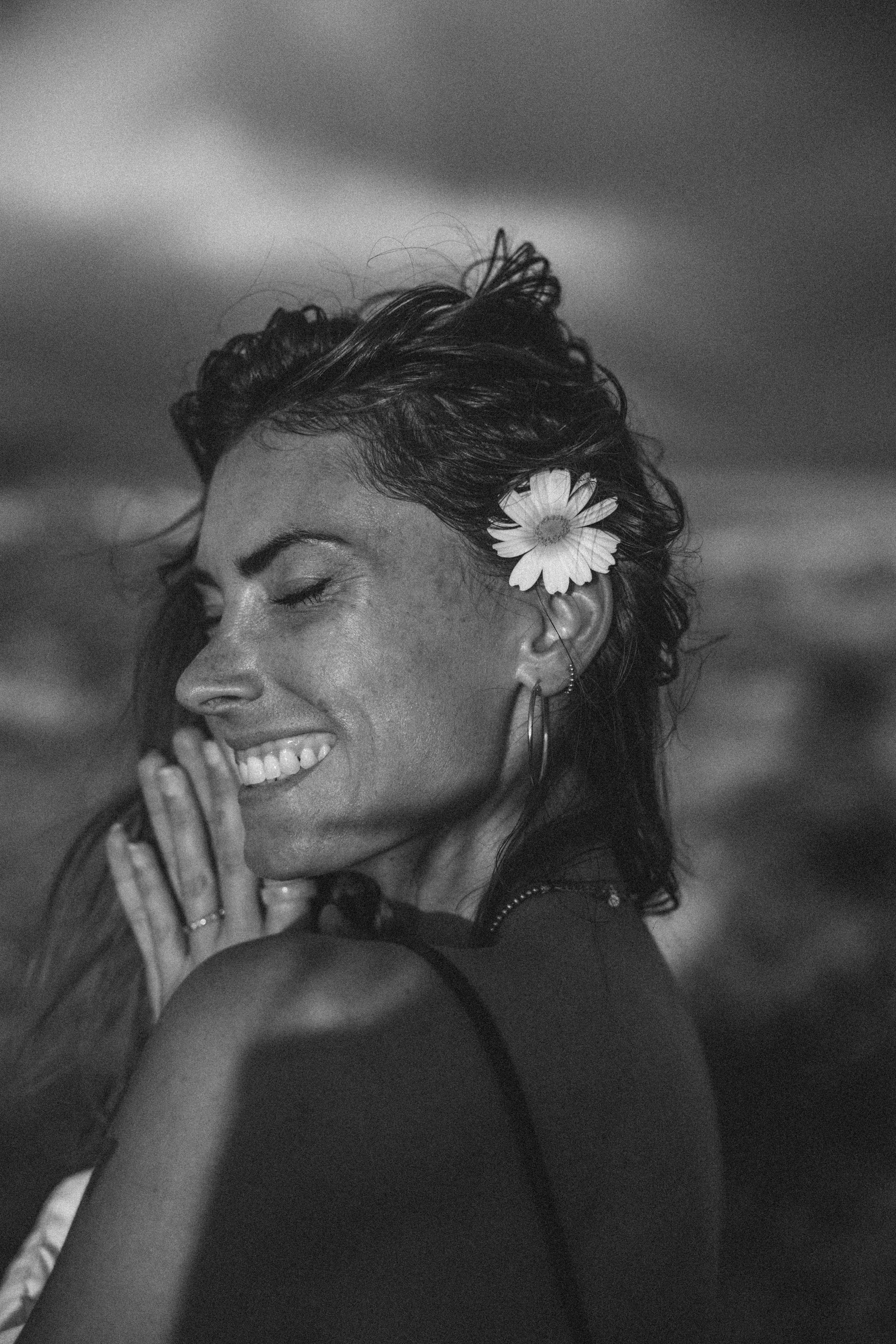 Schwarz-Weiß-Fotografie einer lächelnden Frau mit offener Augenpartie, die einen weißen Blumen in den Haaren trägt und ihre Hände vor dem Gesicht hält.