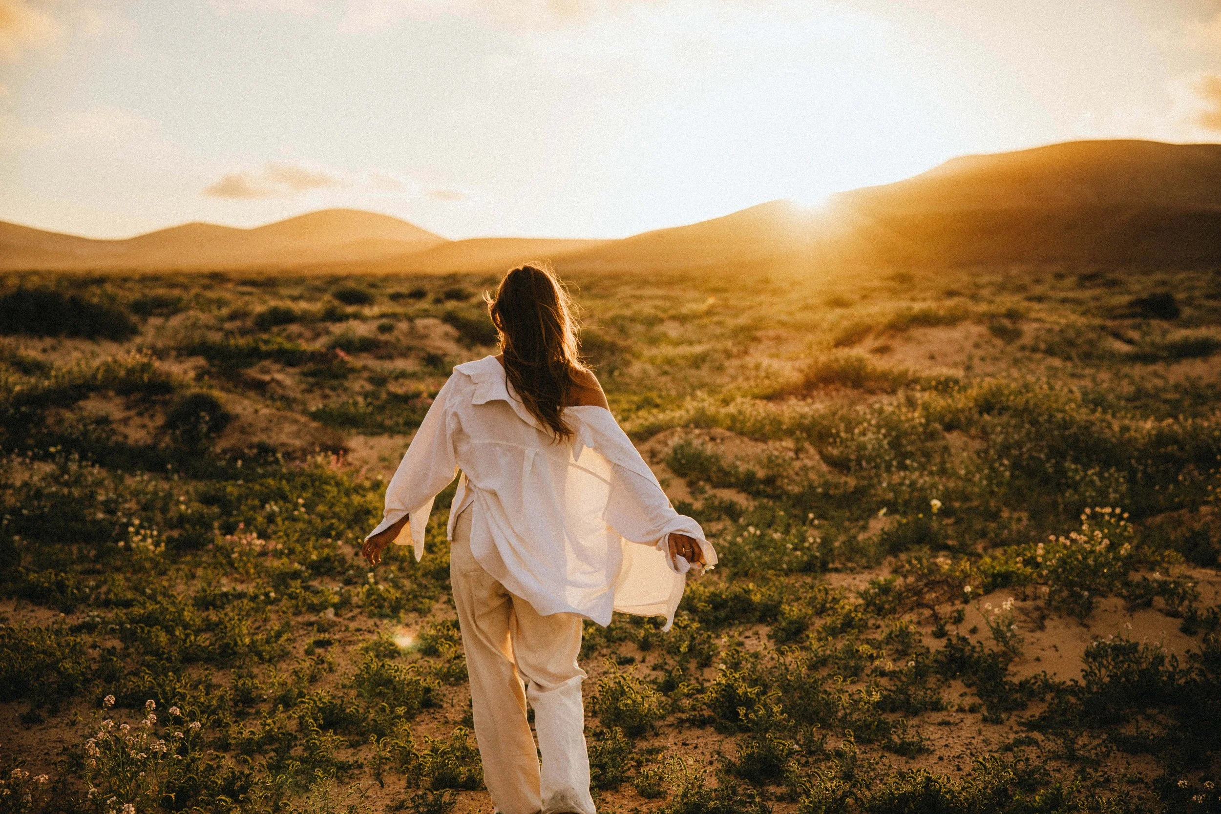 Eine Frau läuft im Sonnenuntergang durch eine Wüstenlandschaft mit kleinen Sträuchern und Hügeln im Hintergrund.