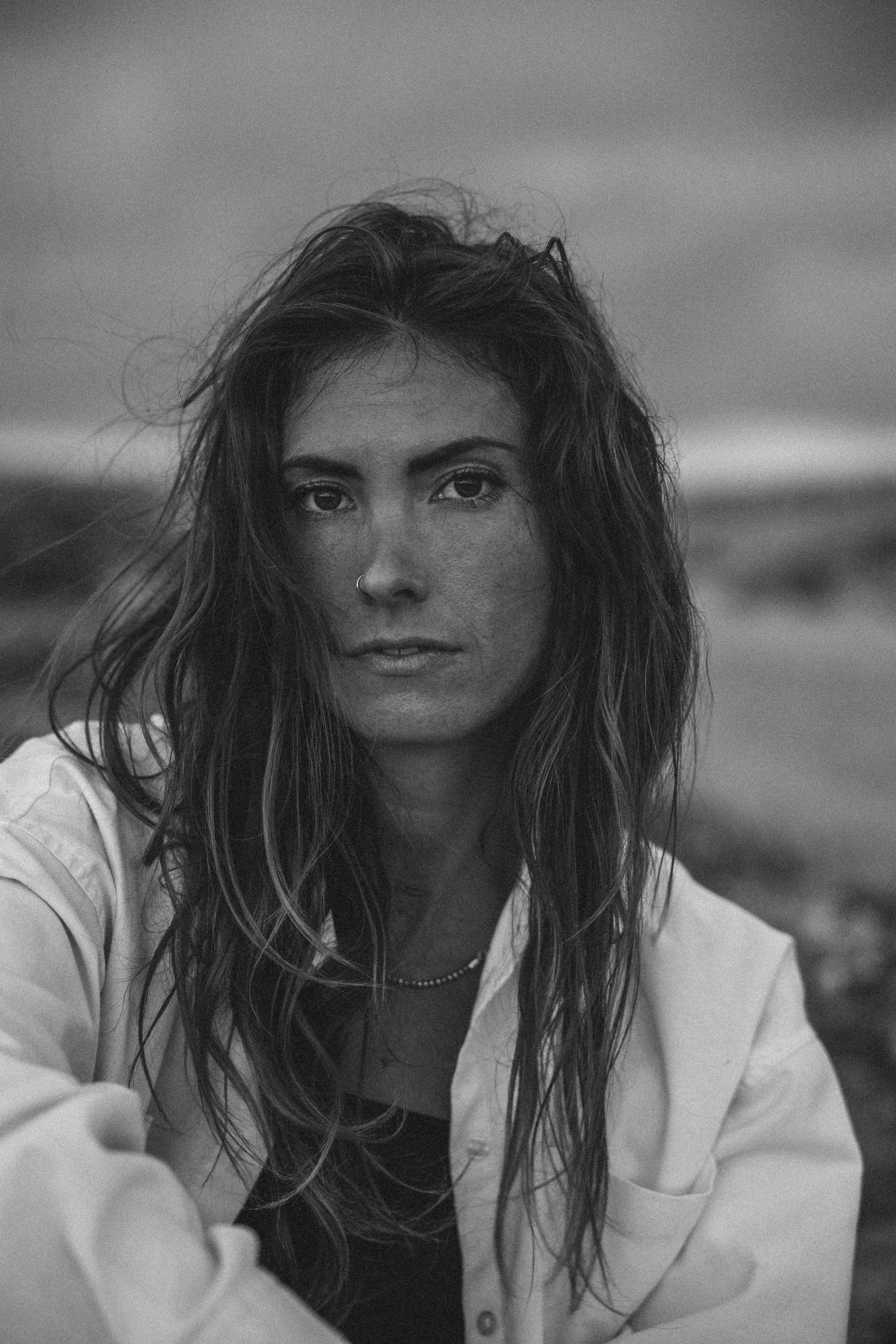 Schwarz-weiß-Foto einer jungen Frau mit langem, wellenförmigem Haar, Nasenring und dunklem Shirt, die draußen vor einer natürlichen Landschaft sitzt.