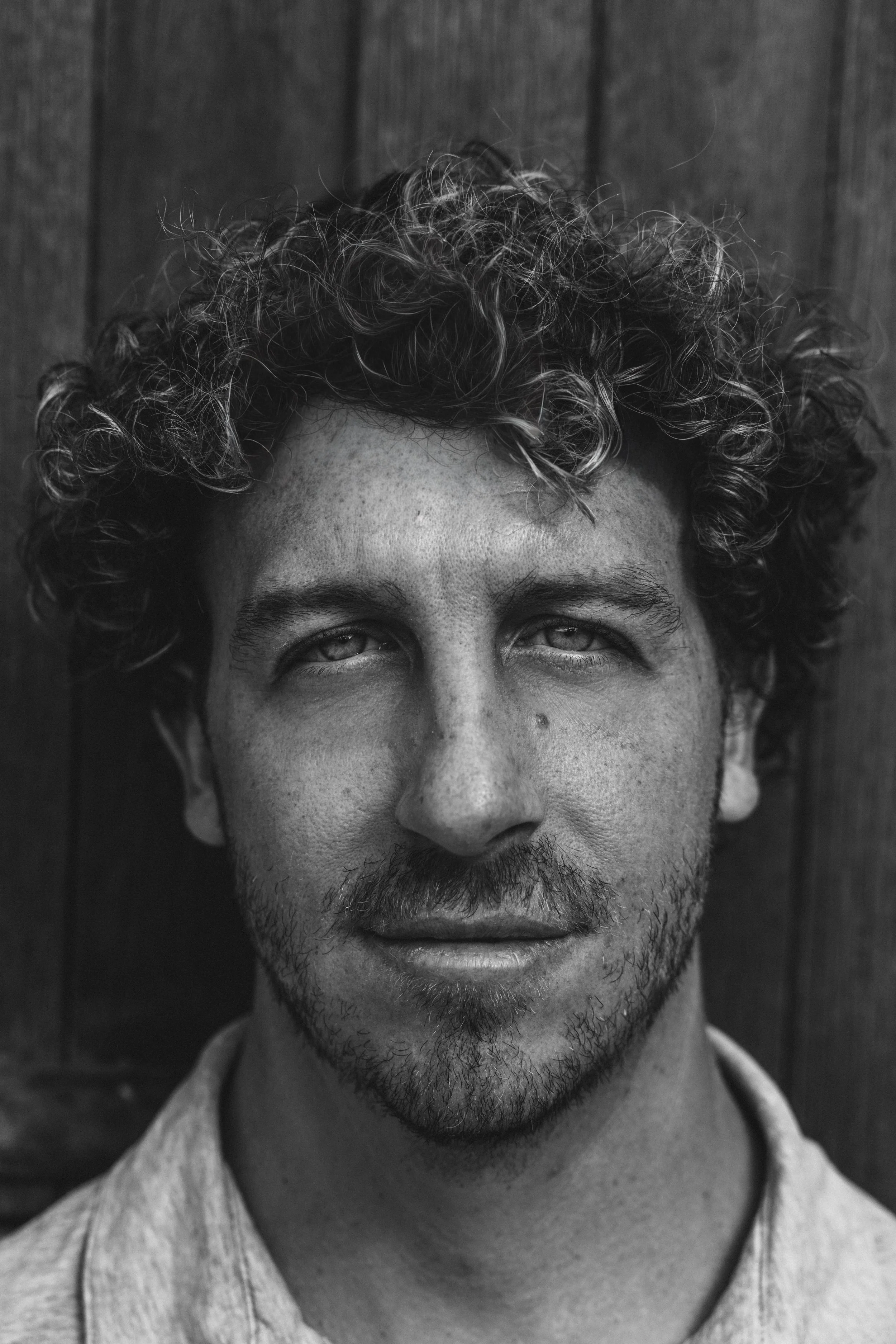 Schwarz-weißer Porträt eines Mannes mit lockigem Haar vor einer Holzvertäfelung.