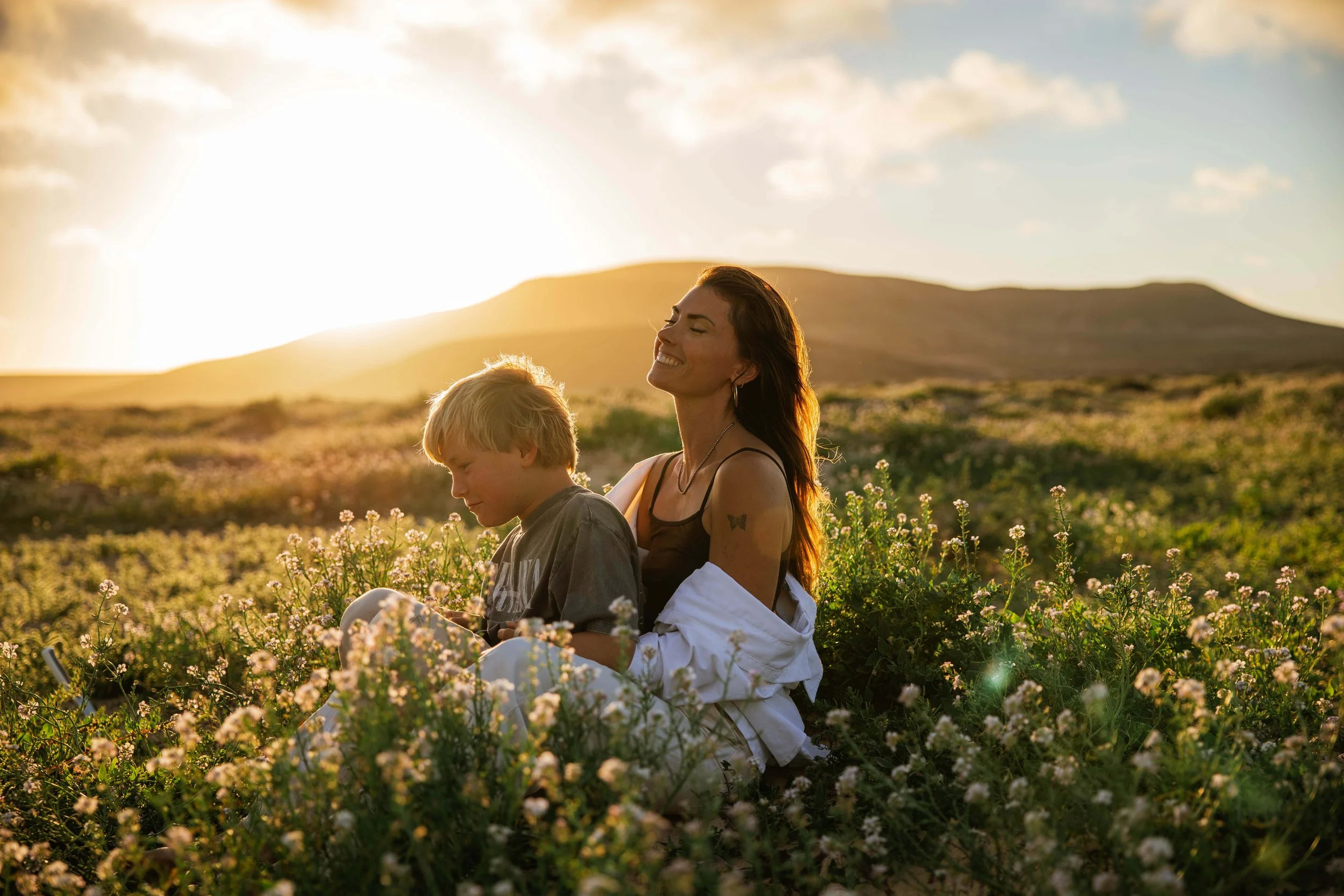 Eine Frau und ein Kind sitzen in einem blühenden Feld bei Sonnenuntergang, die Frau lächelt entspannt