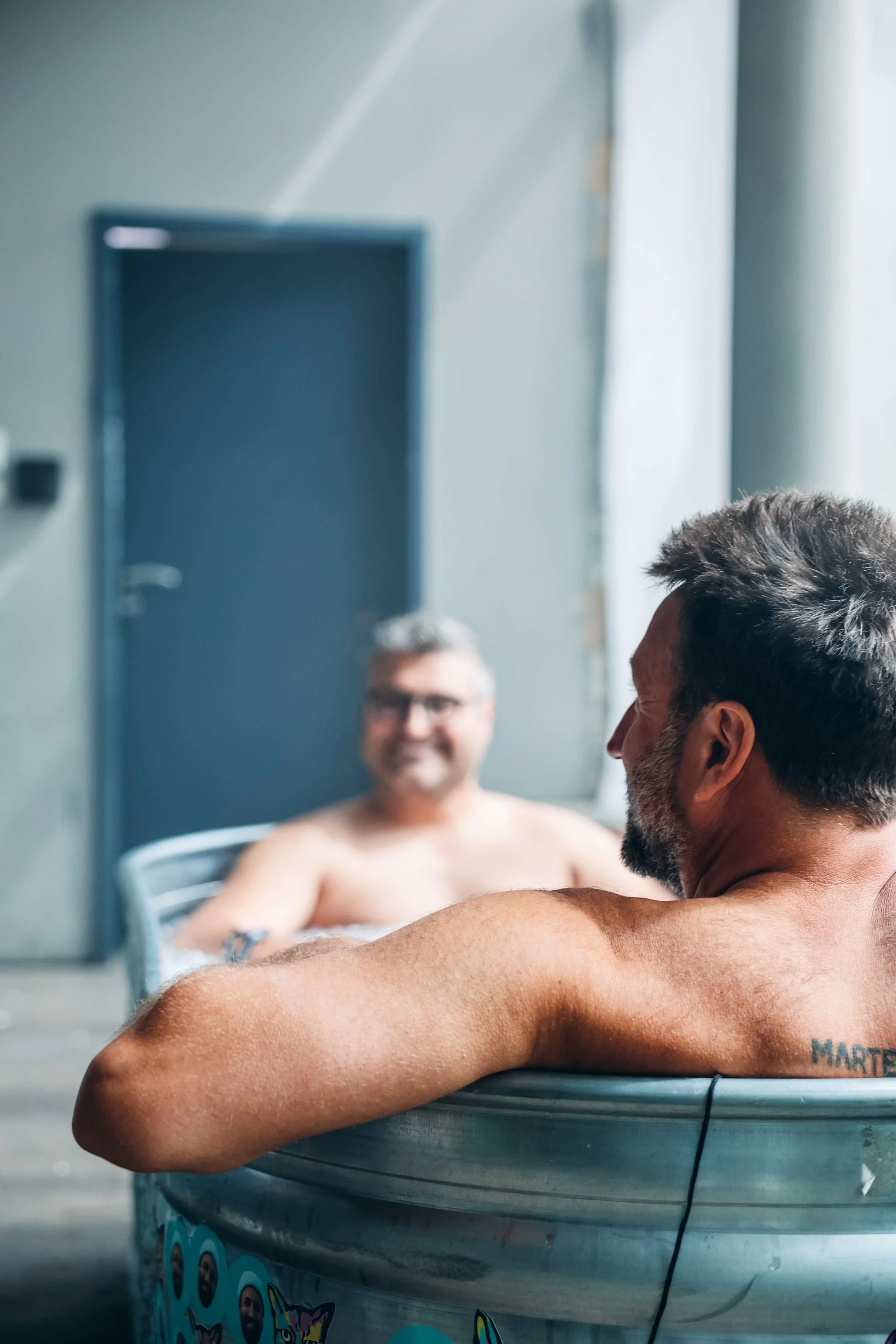 Zwei Mann sitzen entspannt in einer Wanne im Raum, die Wanne ist mit bunten Aufklebern dekoriert und sie haben beide kurze, graue und braune Haare. Einer lächelt in die Kamera, während der andere nur in die Seite schaut.