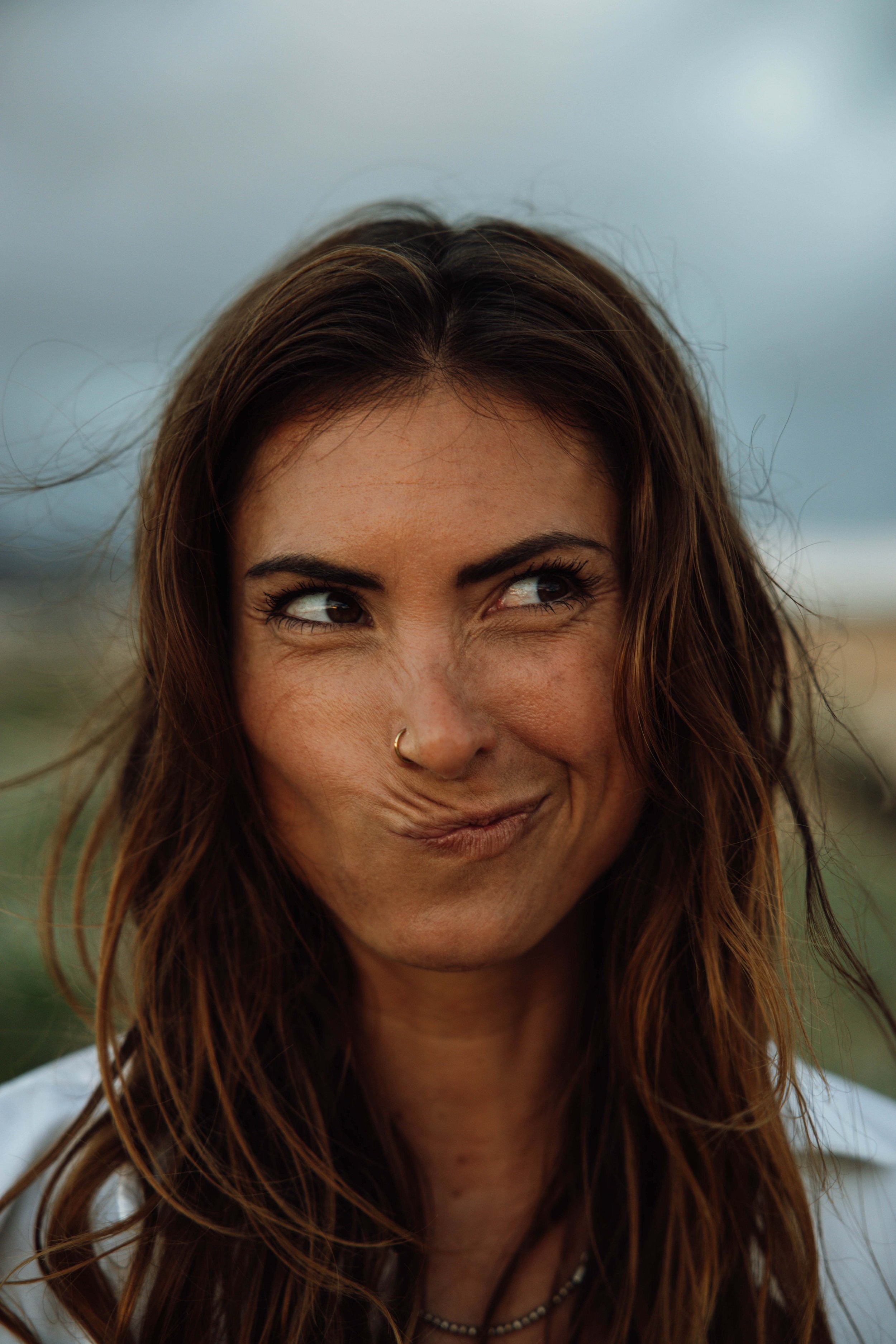 Eine Frau mit braunen, gewellten Haaren macht eine skeptische Gesichtsausdruck, während sie nach rechts schaut, vor einem bewölkten Himmel.