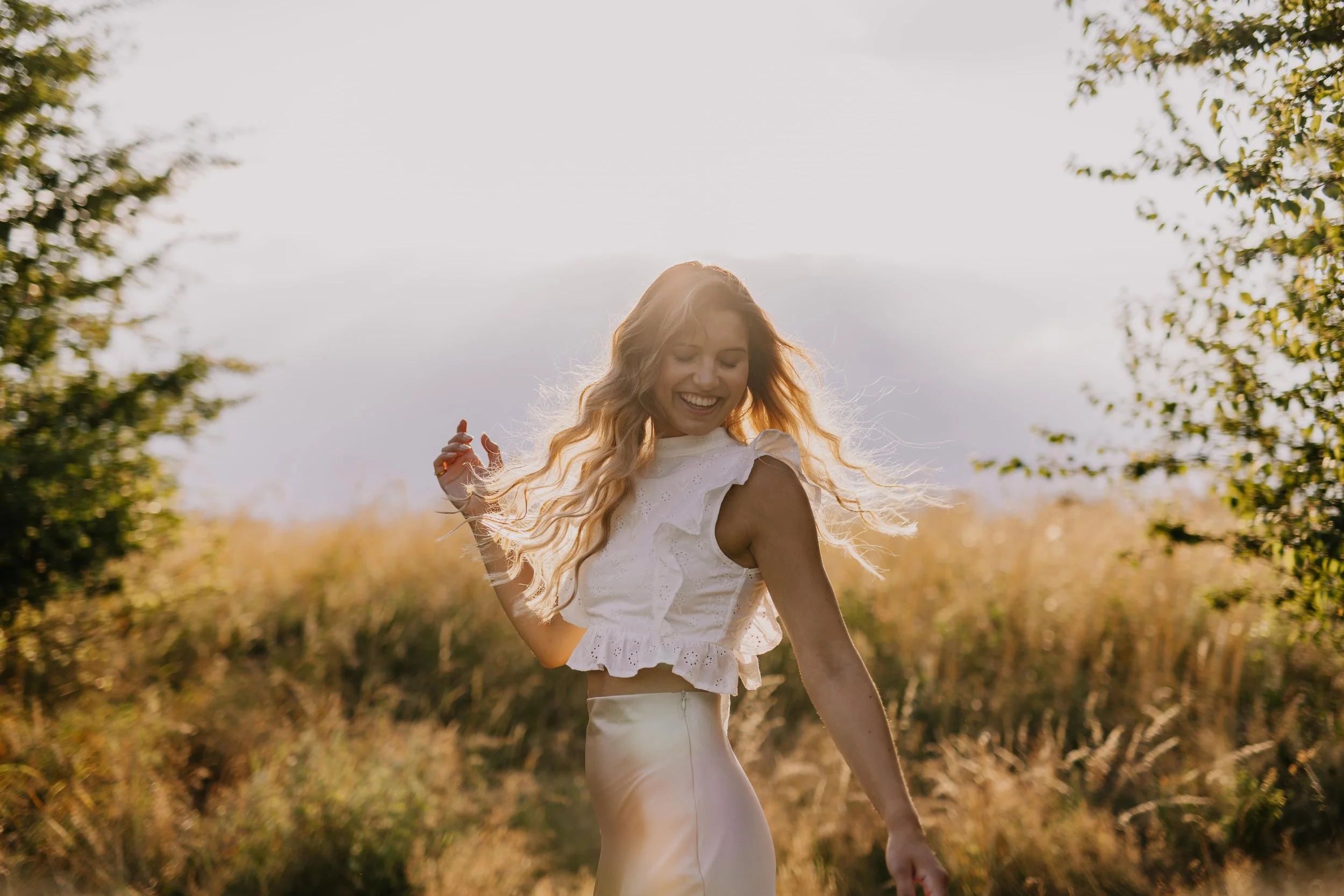 Eine lächelnde Frau mit weichem, gewelltem, blondem Haar tanzt im Sonnenuntergang in einem Feld mit Pflanzen und Bäumen.