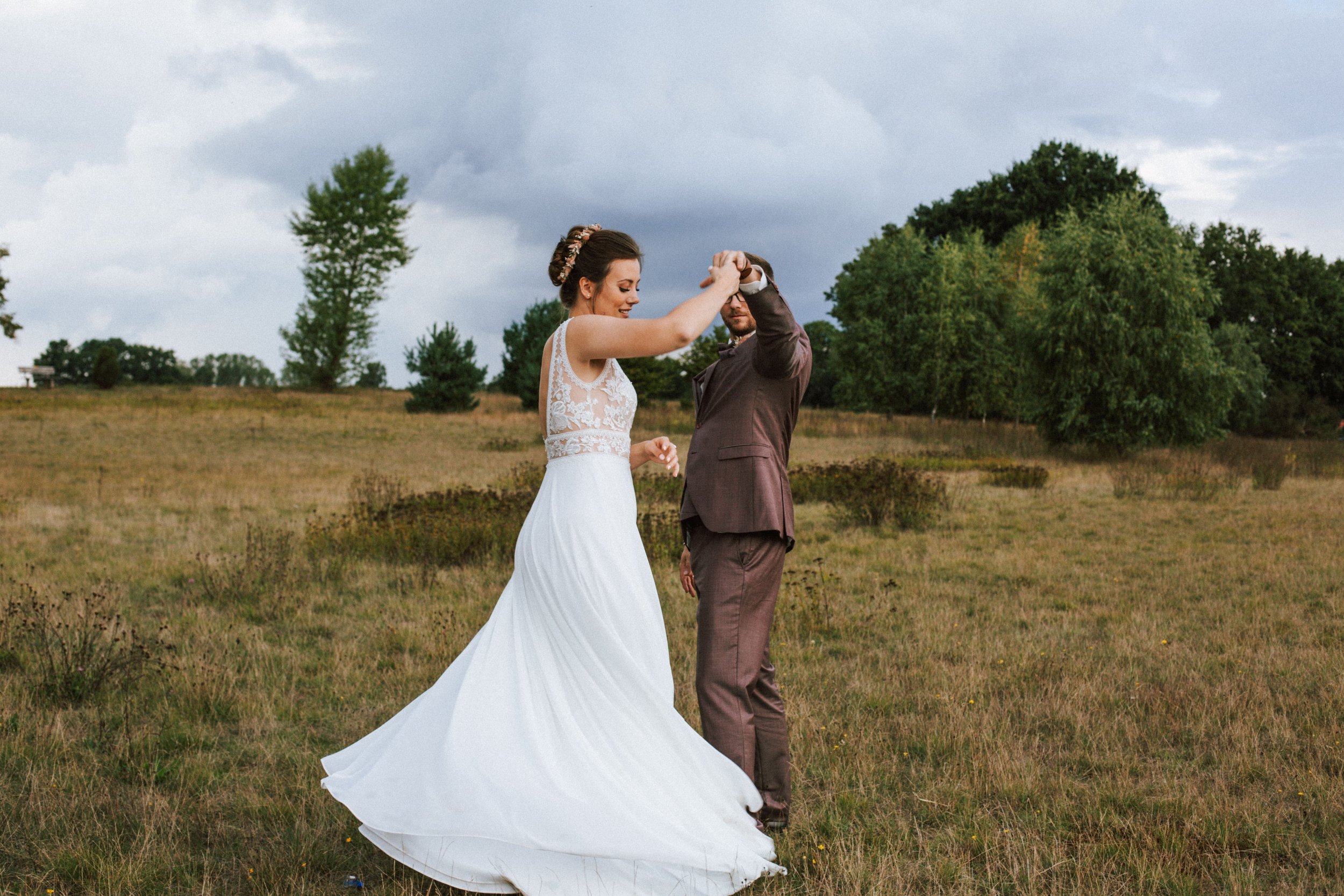 Braut und Bräutigam tanzen im Freien auf einer Wiese bei bewölktem Himmel.